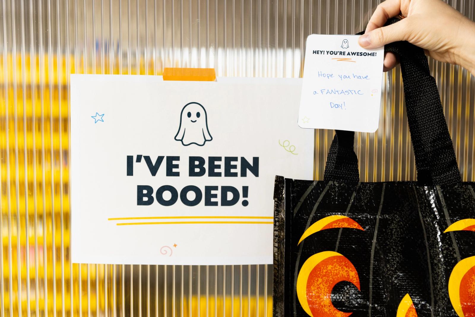 A poster that says "I've been booed" Is taped on a glass door. A hand holds a Halloween bag and small note.