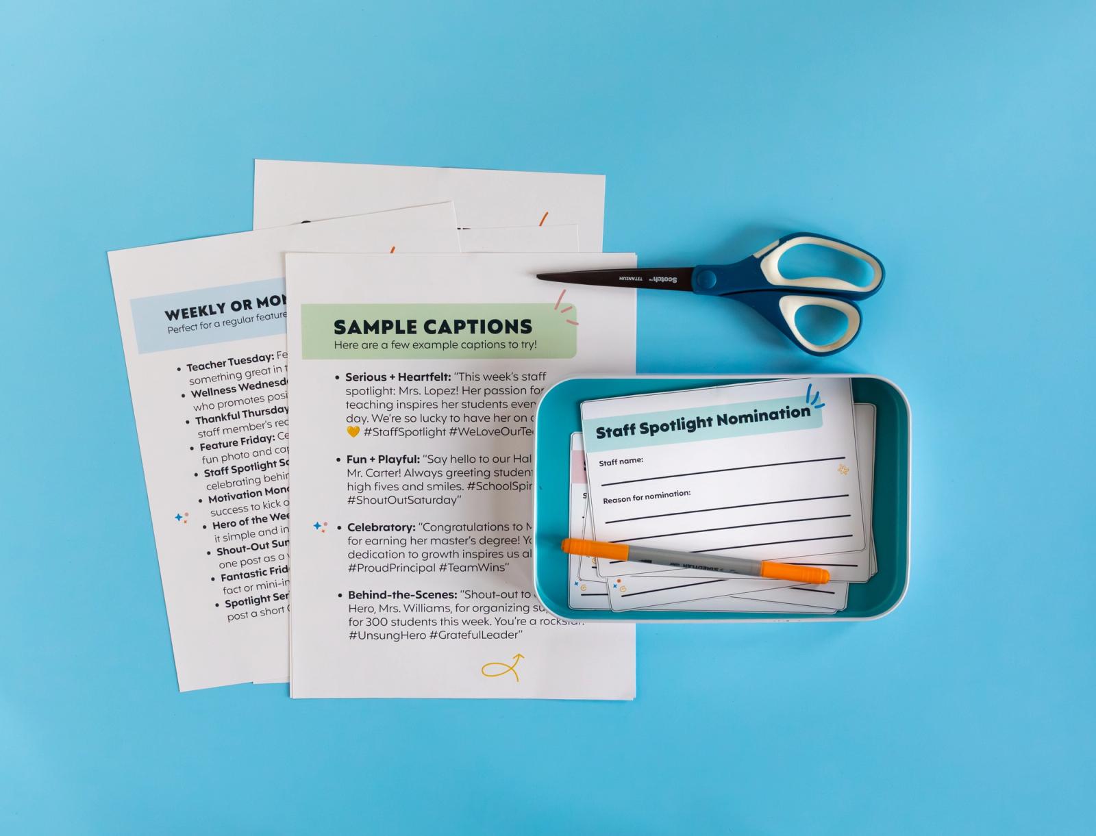Sheets of paper lay on a light blue background. A bin with notecards and a pen in it lays on top of the paper. A pair of scissors is next to the bin. 