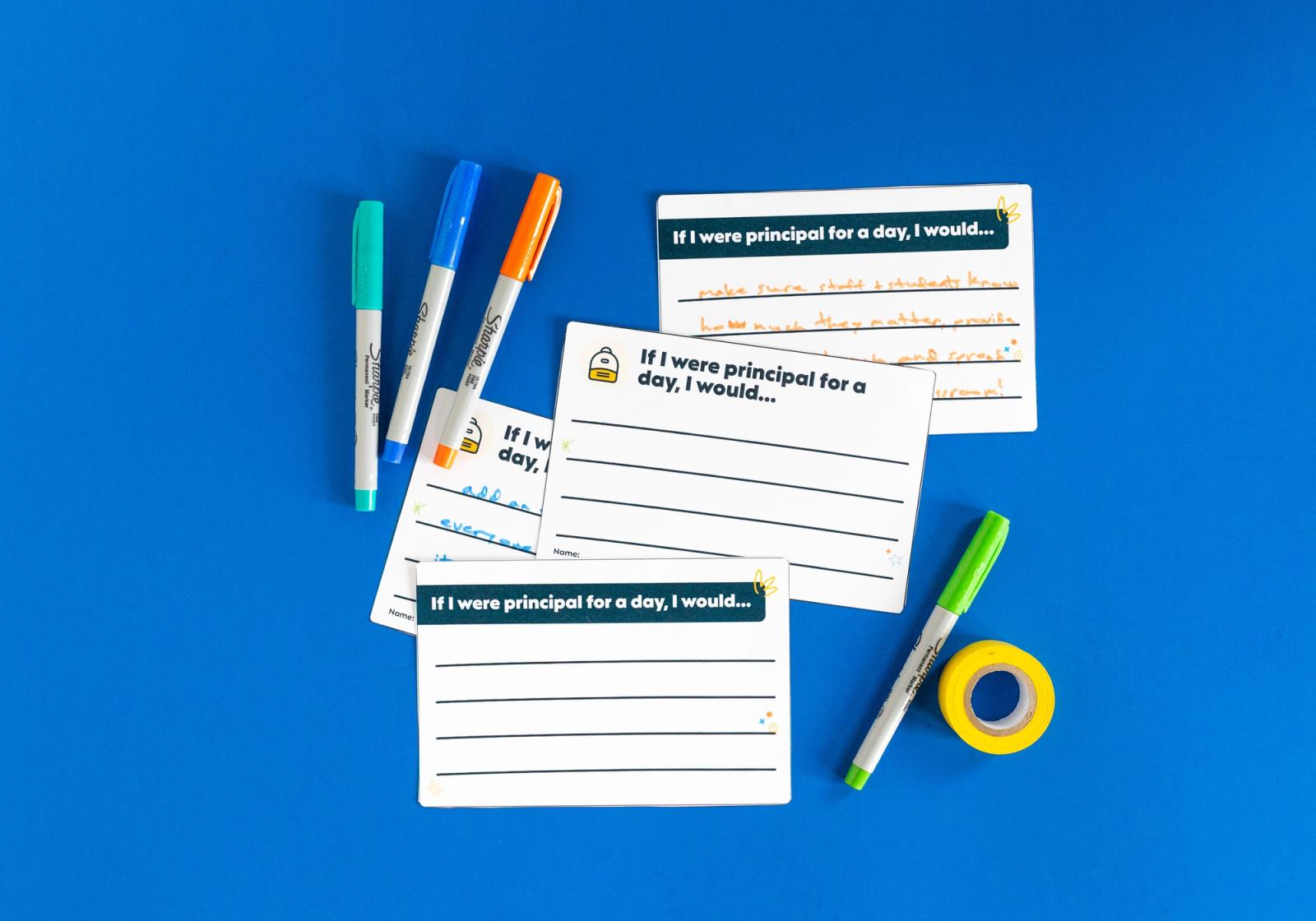 Four notecards with writing on them lay on a blue background. Colorful pens and a roll of tape lay beside them.