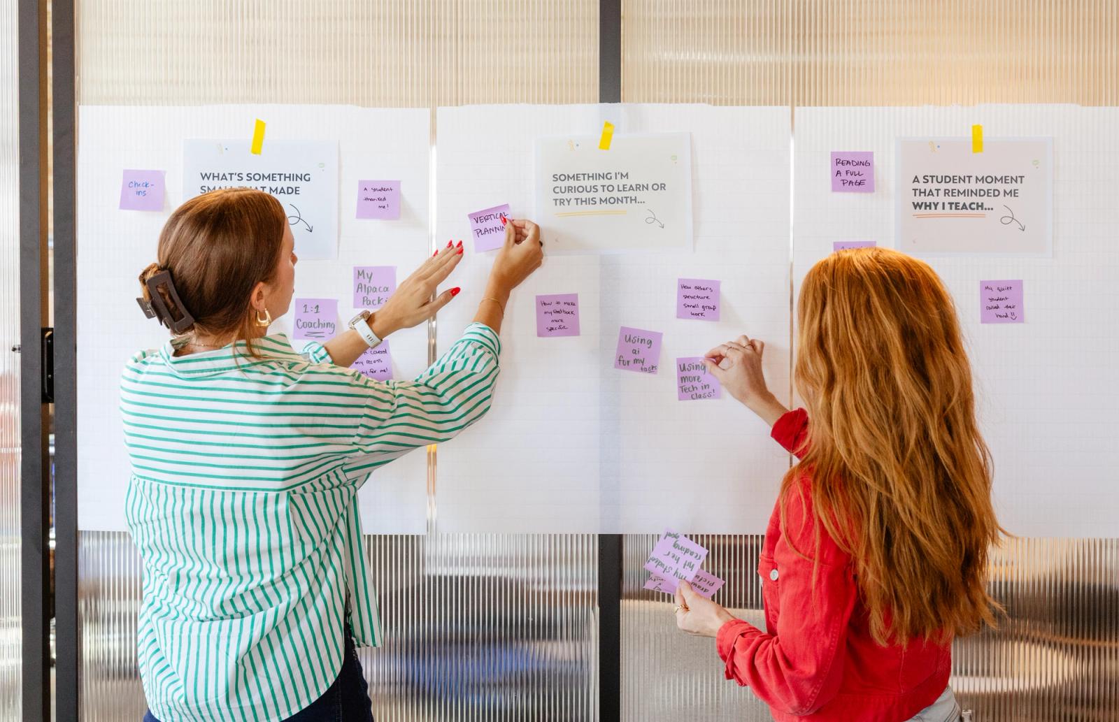 Papers are taped to a white background with purple sticky notes around them. Two women stand and place more notes.