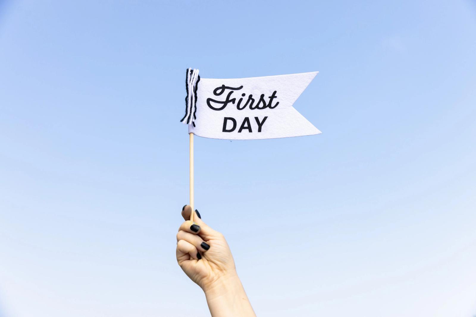A hand holds up a pennant that says "first day" on it. Blue sky in the background.