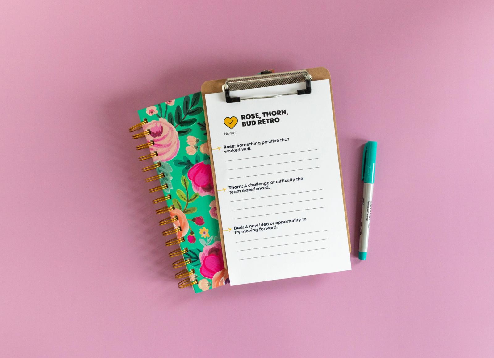 A clipboard with a piece of paper that says "rose, bud, thorn retro" on it lays on a floral notebook on a light purple background. A blue pen lays next to the notebook.