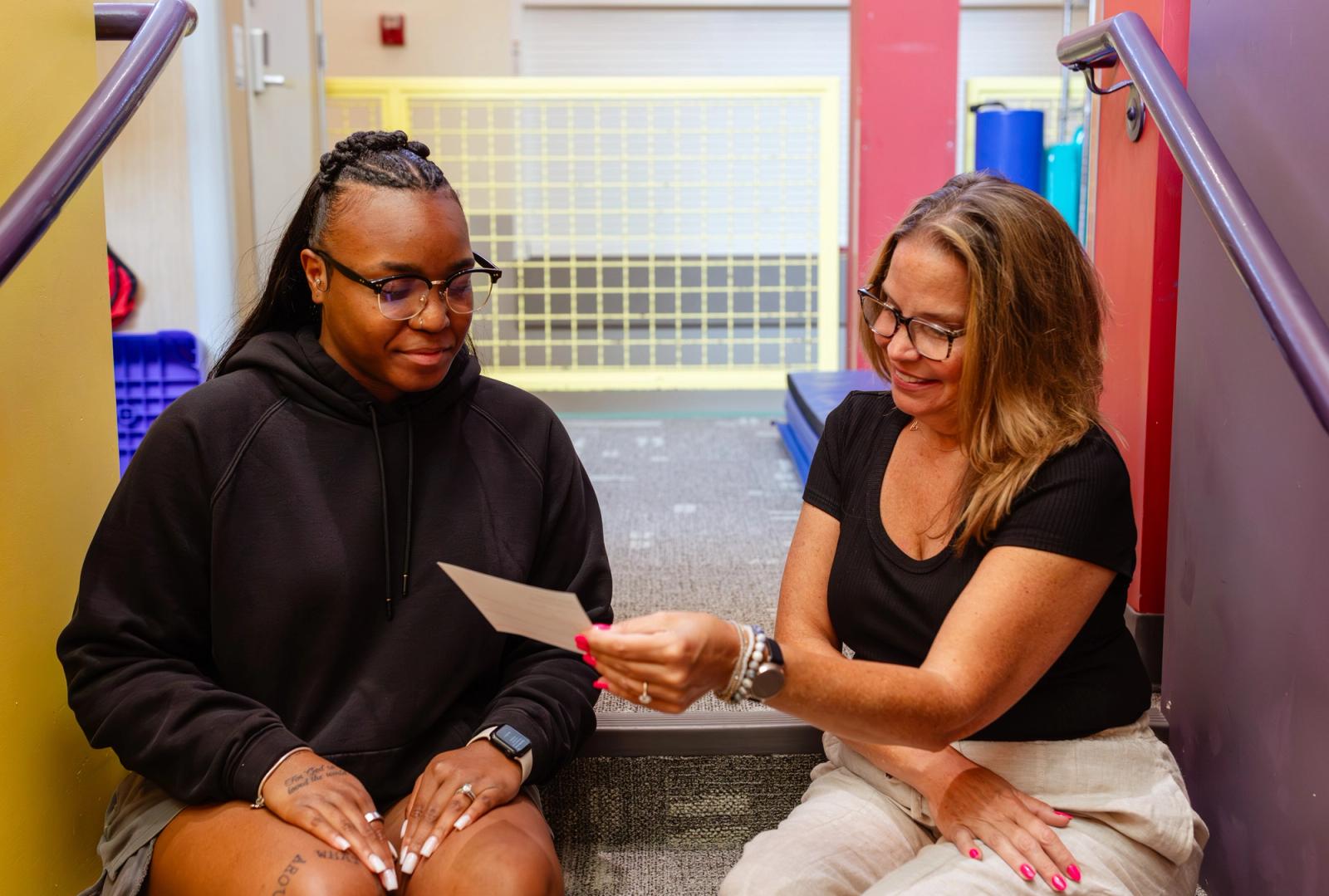 Two women sit in a stairwell look at a notecard that is between them.