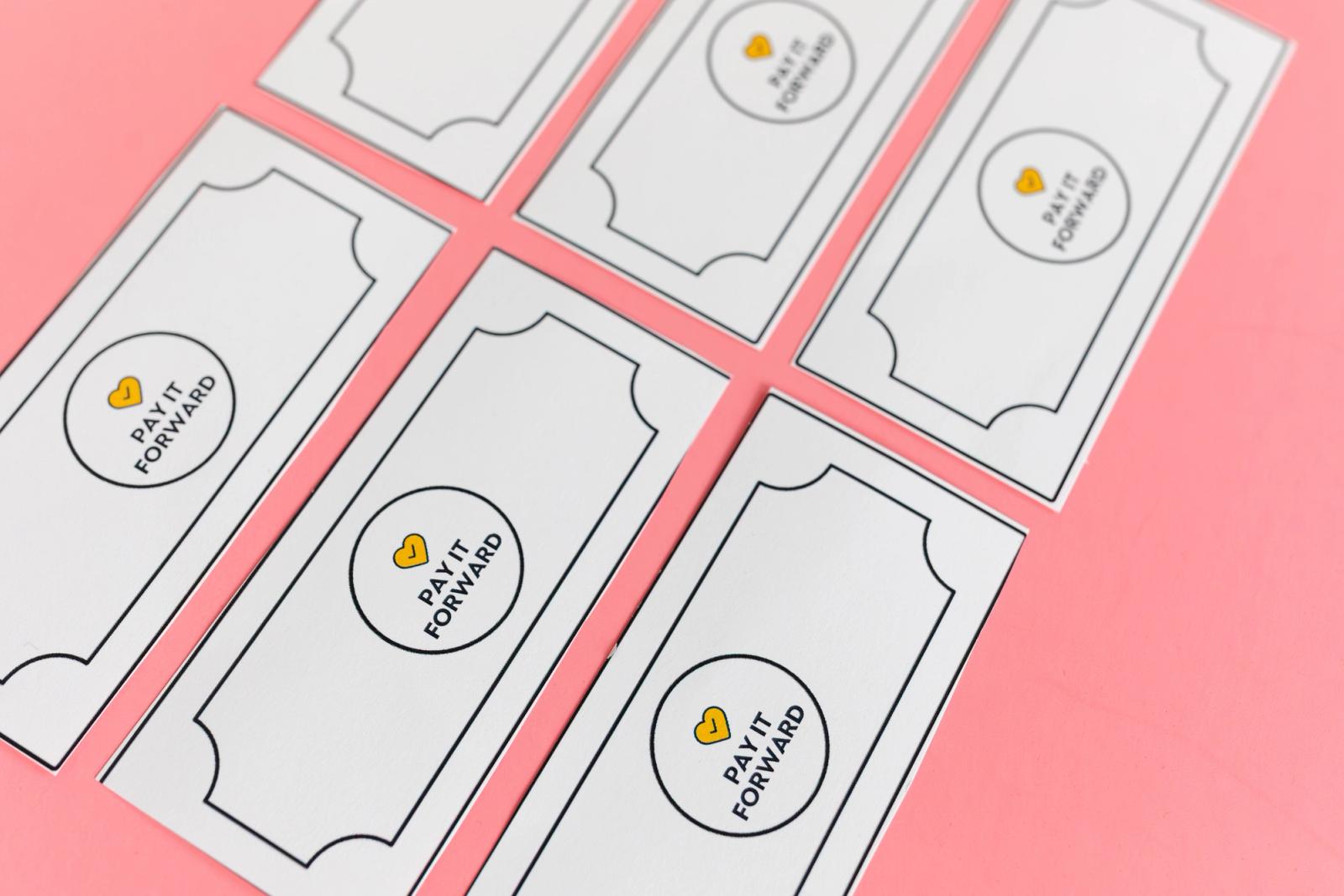 Six coupons that says "pay it forward" lay diagonally on a salmon background.