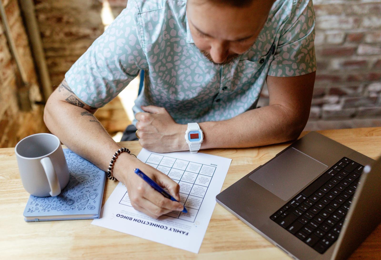 A man sits at a desk with a notebook, coffee cup, and open laptop. He fills in a bingo card with a blue pen.