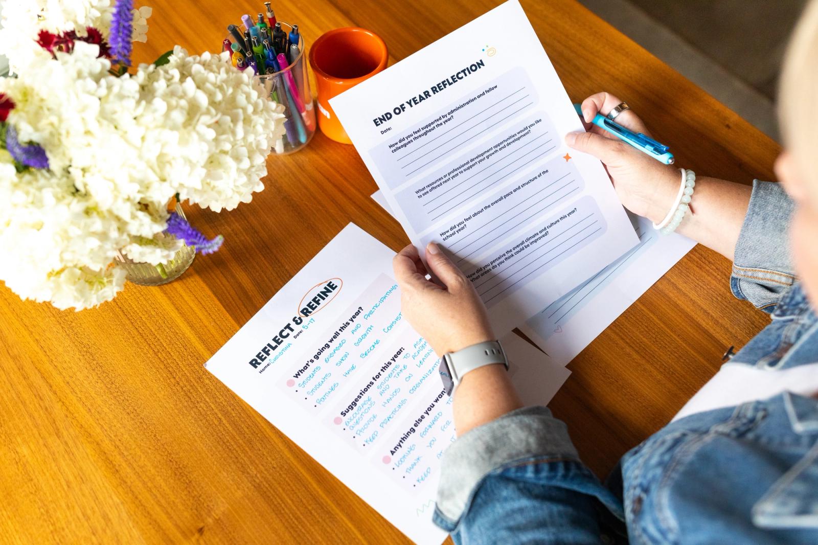 A woman sits at a wood desk with white flowers, pens, and an orange coffee mug on it. She holds a piece of paper in her hands with more papers on the desk.