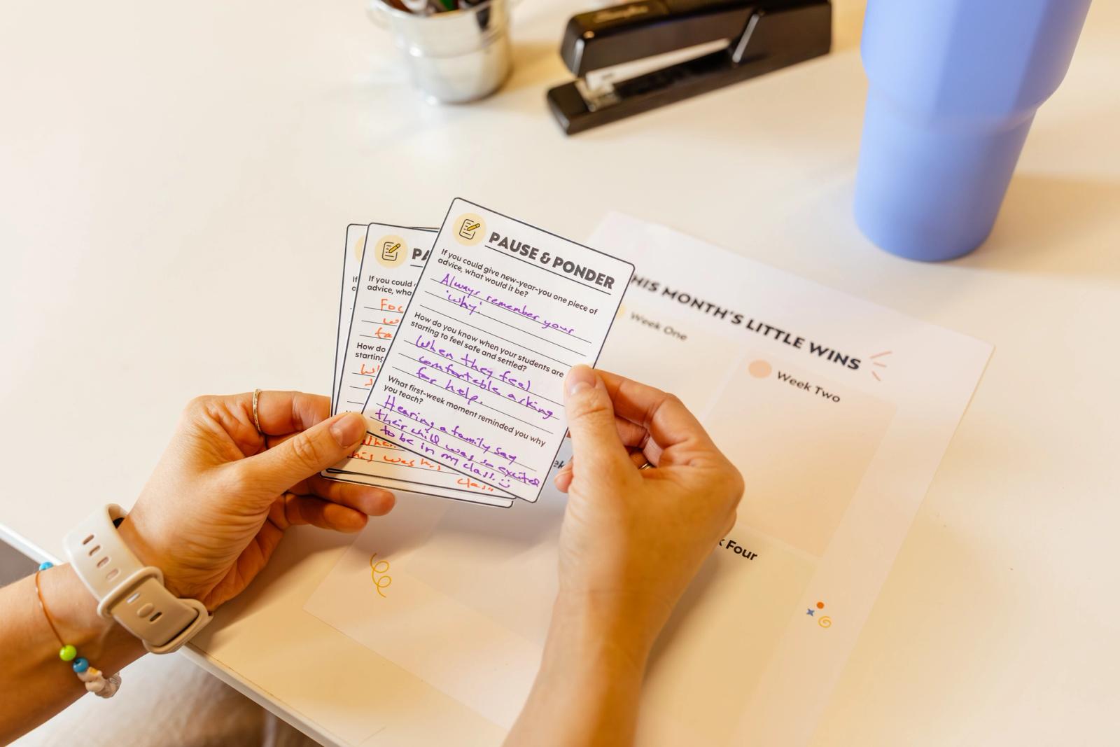 Two hands shuffle through notecards with written message on them. Background is a white table with a blue water bottle and stapler on it.