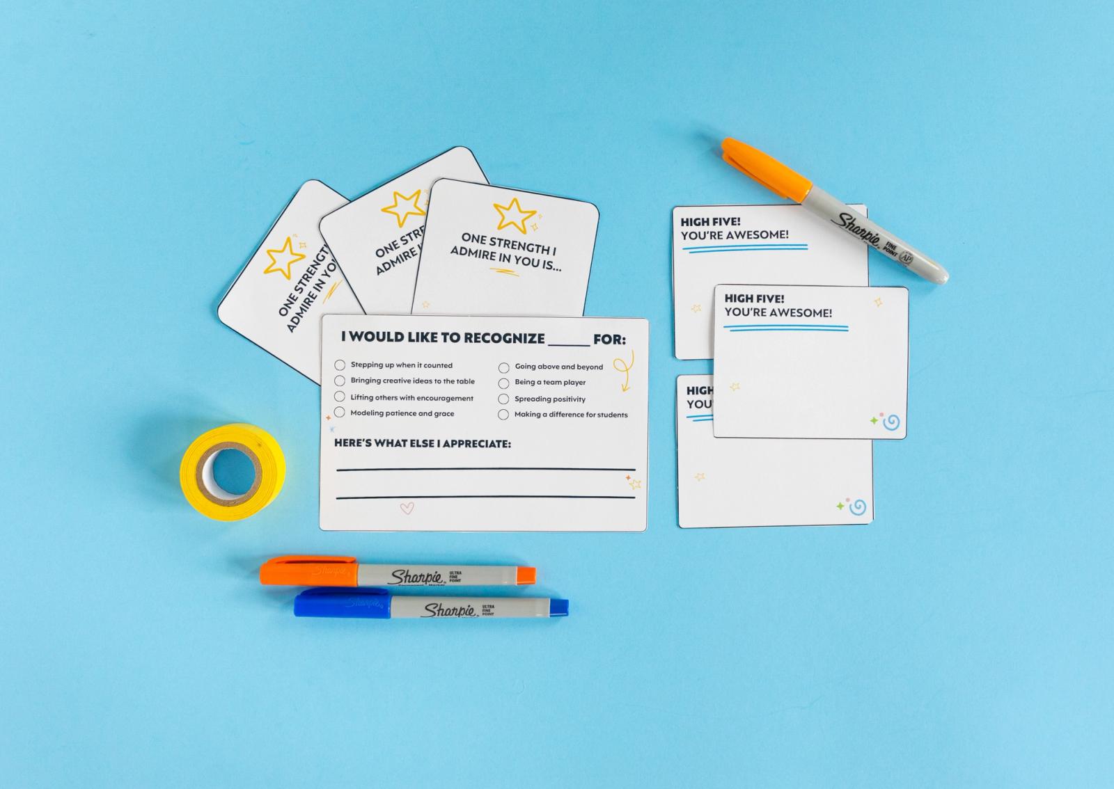 Small note cards, orange and blue pens, a yellow roll of tape lay on a light blue background.