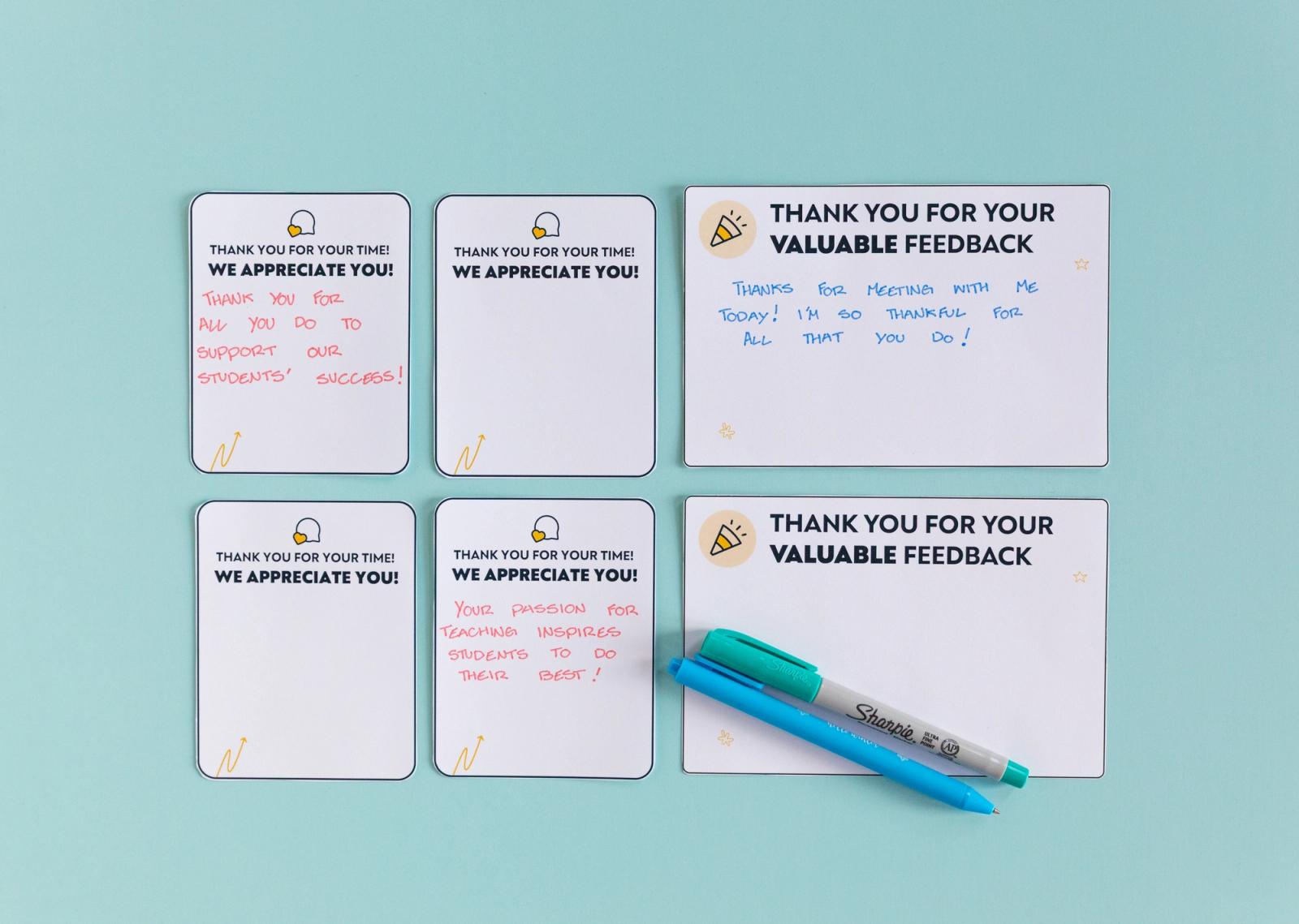 Four small notecards and two larger notecards with writing on them lay on a light blue background with two blue pens on top of them.