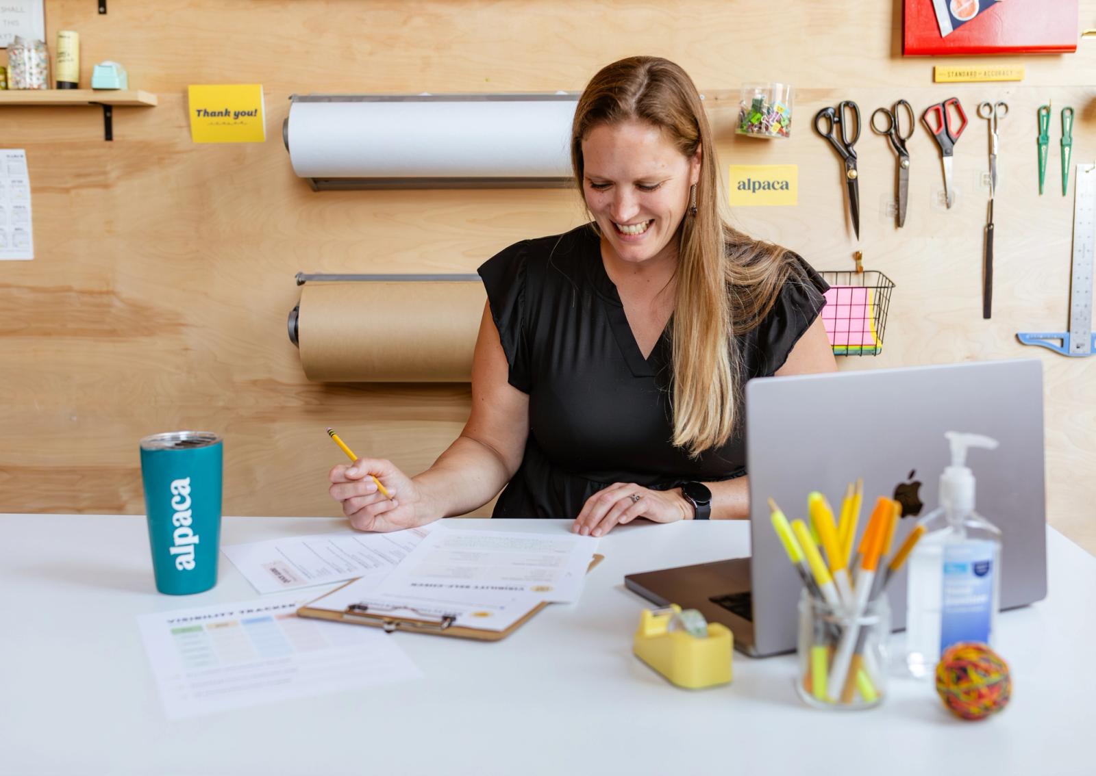 A woman in a black dress sits at a desk and fills out paperwork. A laptop, school supplies, and a coffee cup sit on the desk.