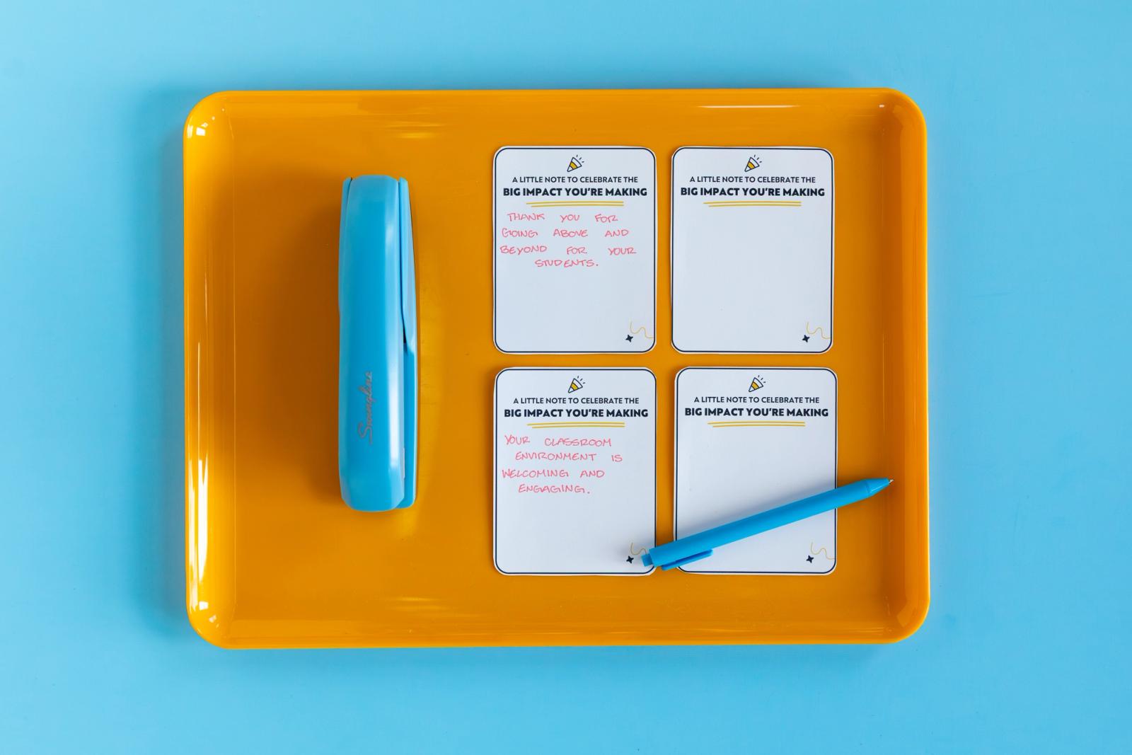 Four small pieces of paper, a blue pen, and blue stapler lay on an orange tray on a blue background.