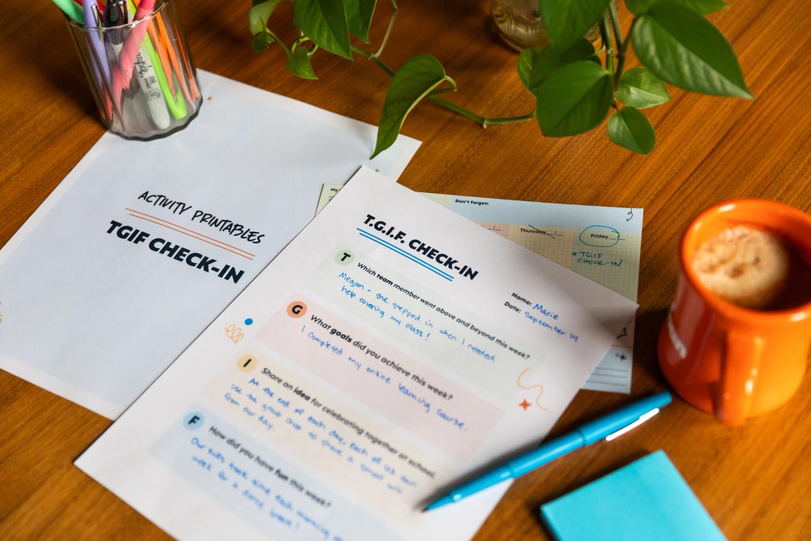 Several sheets of paper, one with writing on it, sit on a wooden desk. A blue pen, sticky notes, jar of pens, small plant, and coffee cup are placed around the paper. 