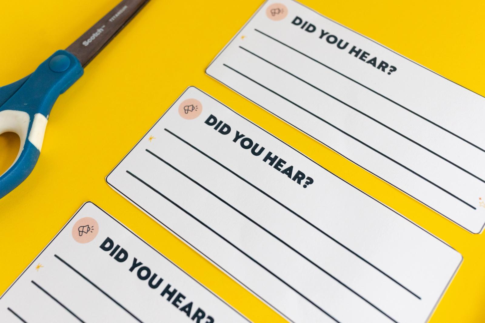 Notecards that say "did you hear" lay diagonally on a yellow background with a pair of blu scissors beside them.