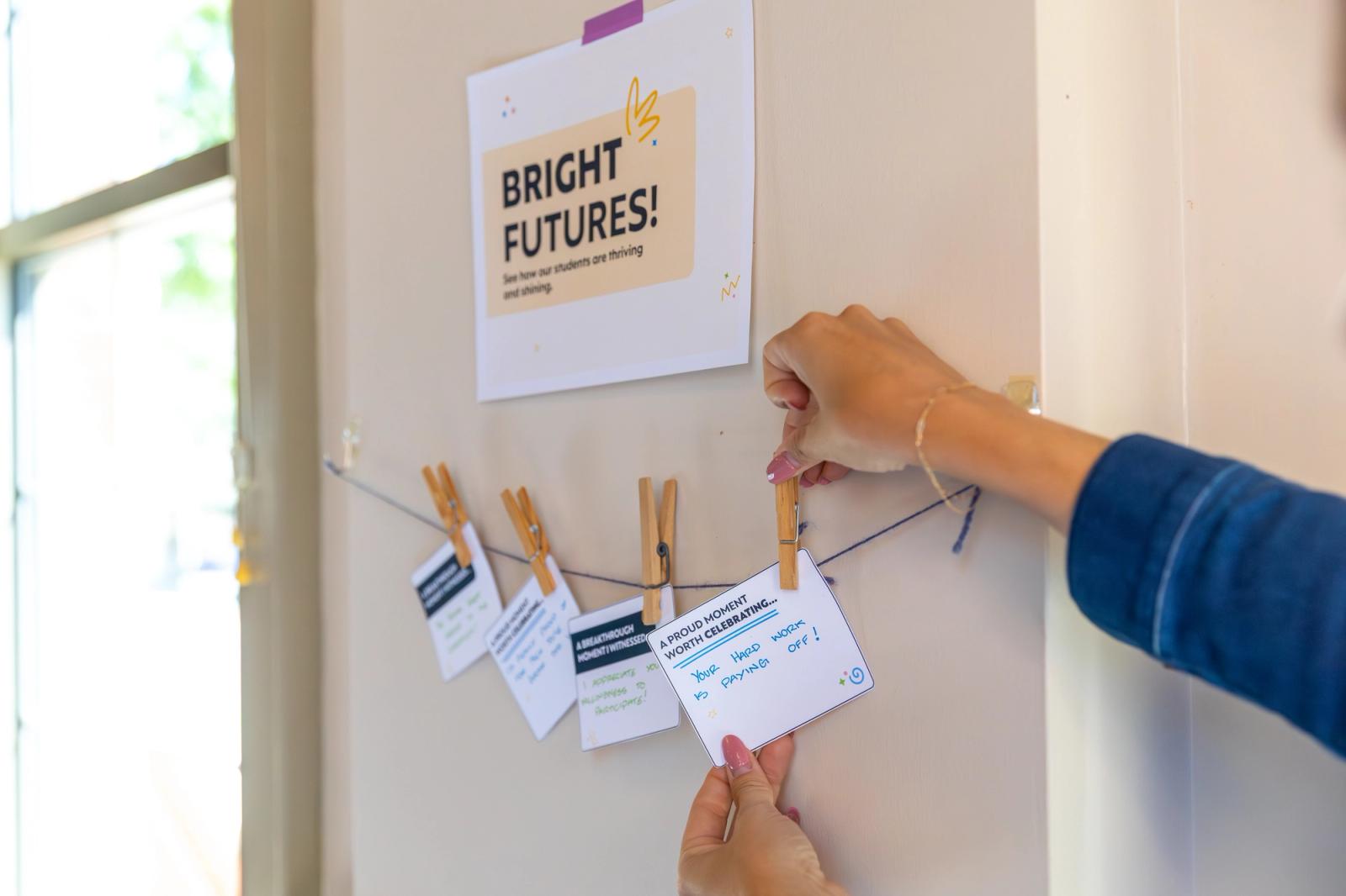 A piece of paper with "bright futures" typed on it hangs on a white wall next to a window. Below is a string with small pieces of paper hanging on it with clothespins. A hand reaches in from the right side to hang one more piece of paper.