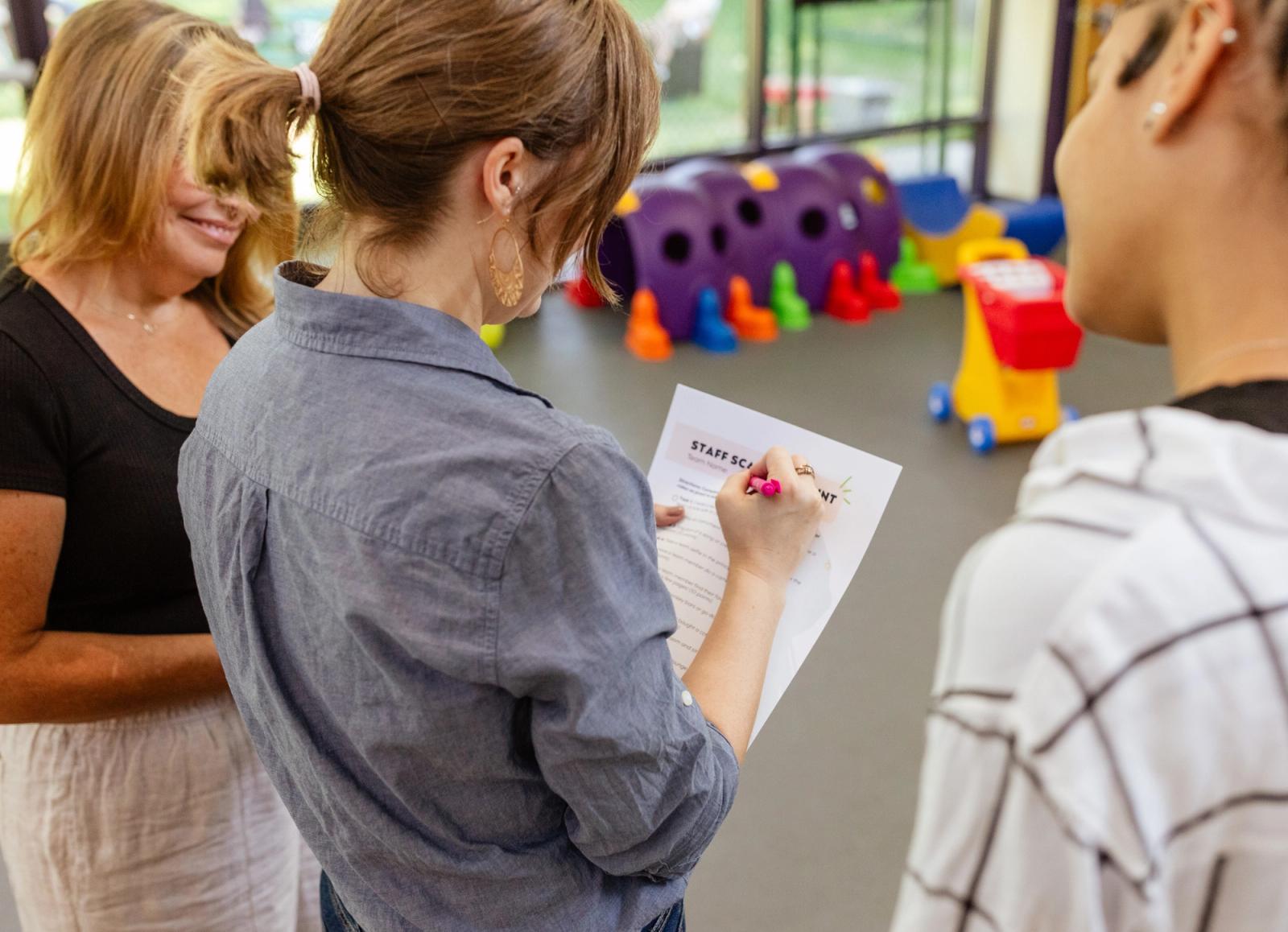Three women stand in a childcare room. One holds a sheet of paper and writes on it while the other two look on.
