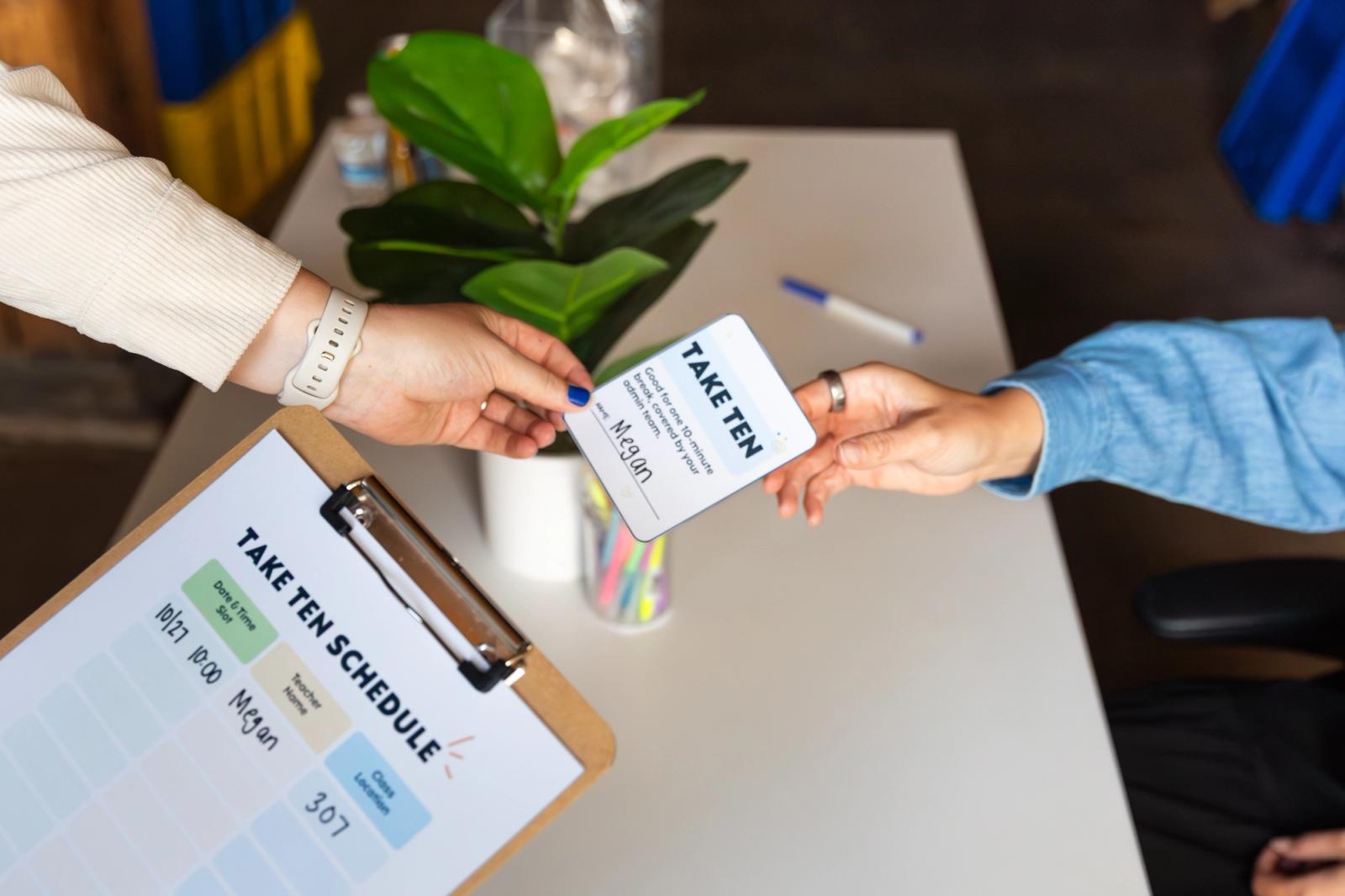 Someone passes a coupon on to another person in front of a white desk with a plan and pens on it.
