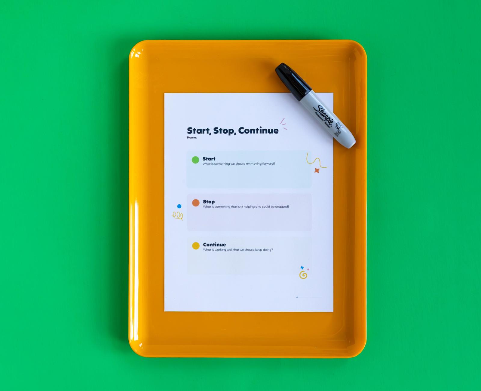 A piece of paper with "start, stop, continue" written on it lays on a yellow tray with a black marker, all on a green background.