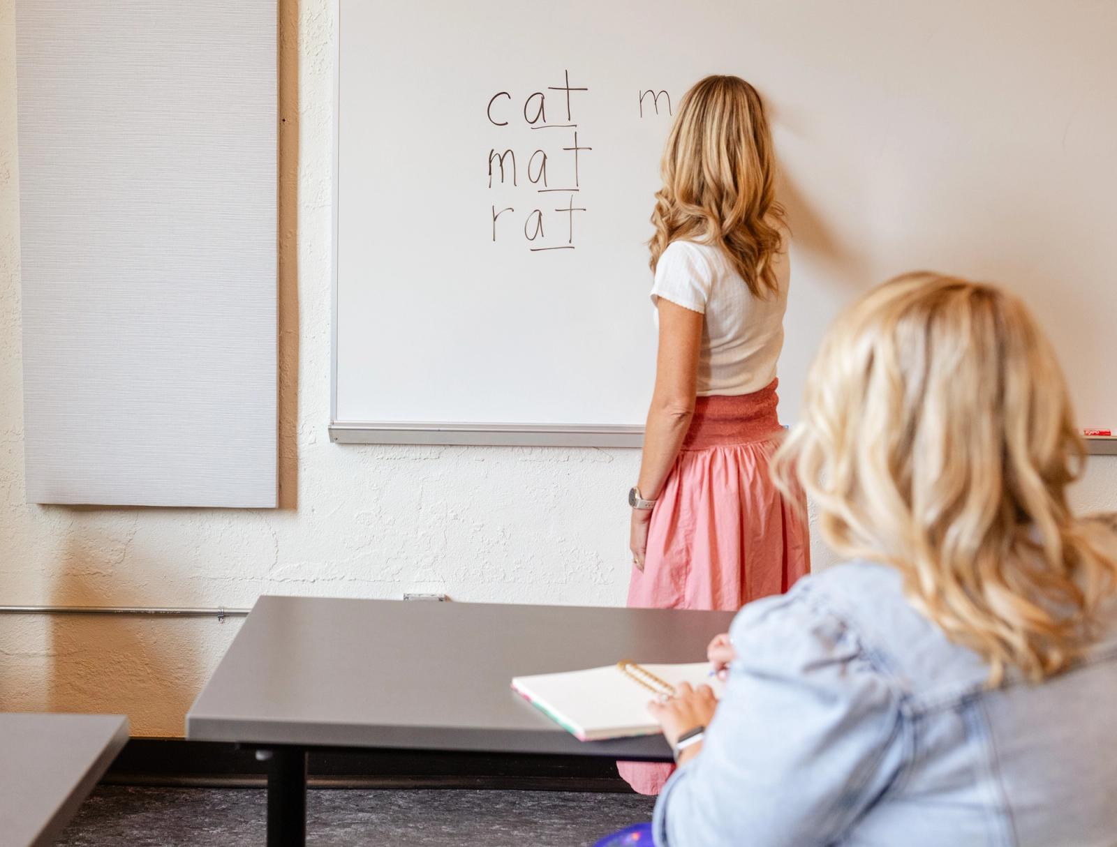 A woman stands writing at a whiteboard. Another woman sits at a table and makes notes in a small notebook.