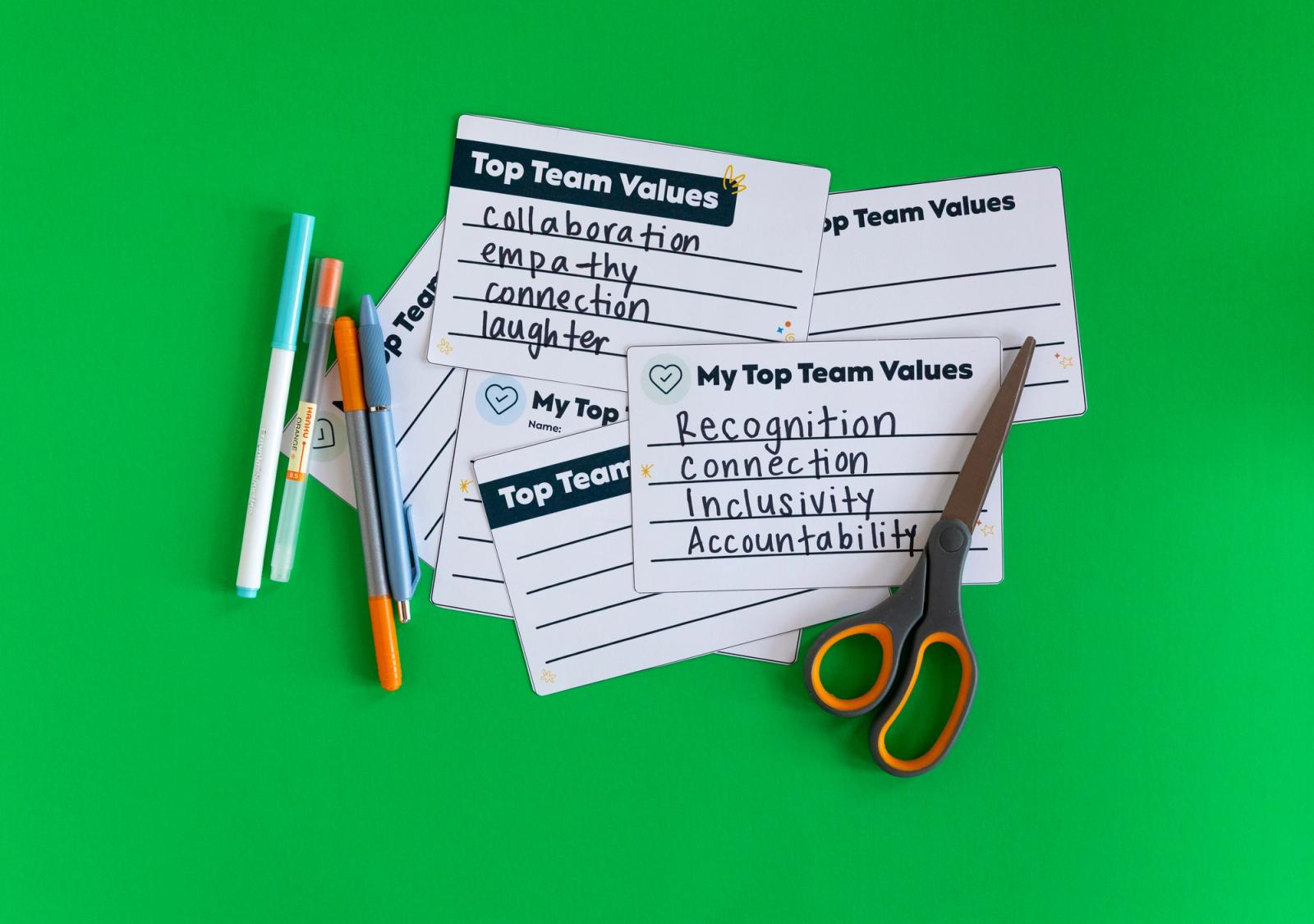 A pile of notecards, pens and a pair of scissors lay on a green background.
