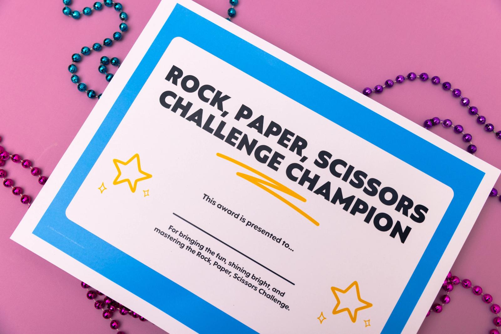 A piece of paper that says :rock, paper, scissors challenge champion" and bead necklaces lay on a pink background.