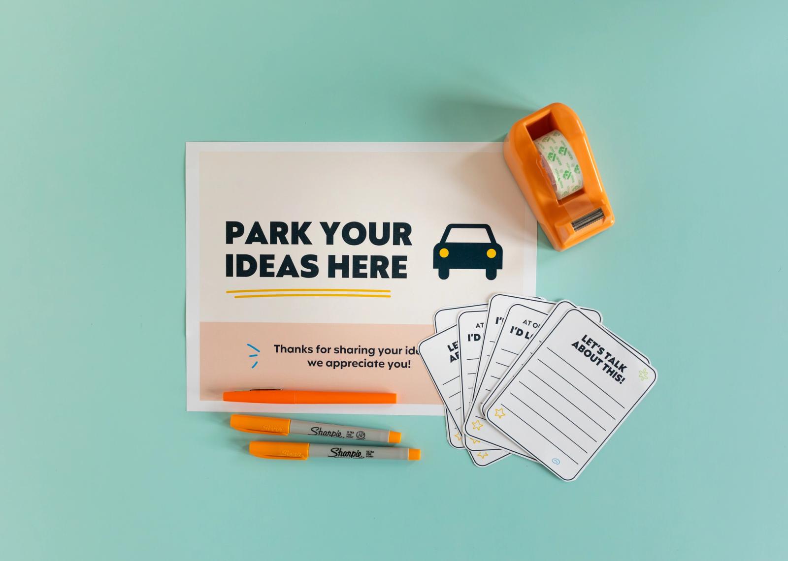 A poster that says "park your ideas here" lays on a light blue background. A tape dispenser, orange pens, and blank notecards lay around the poster.