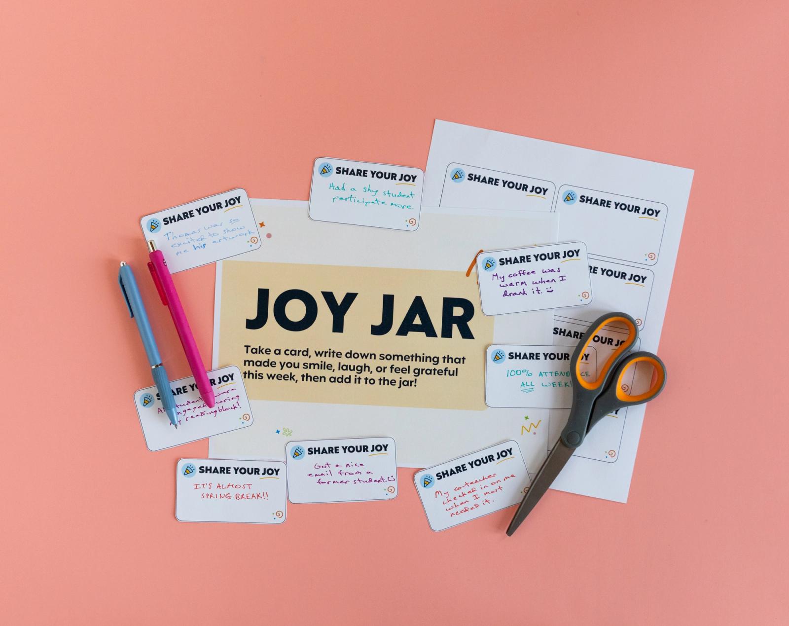A poster that says "joy jar" lays on a salmon background with handwritten notes scattered around it. A pair of scissors and two pens sit beside them.