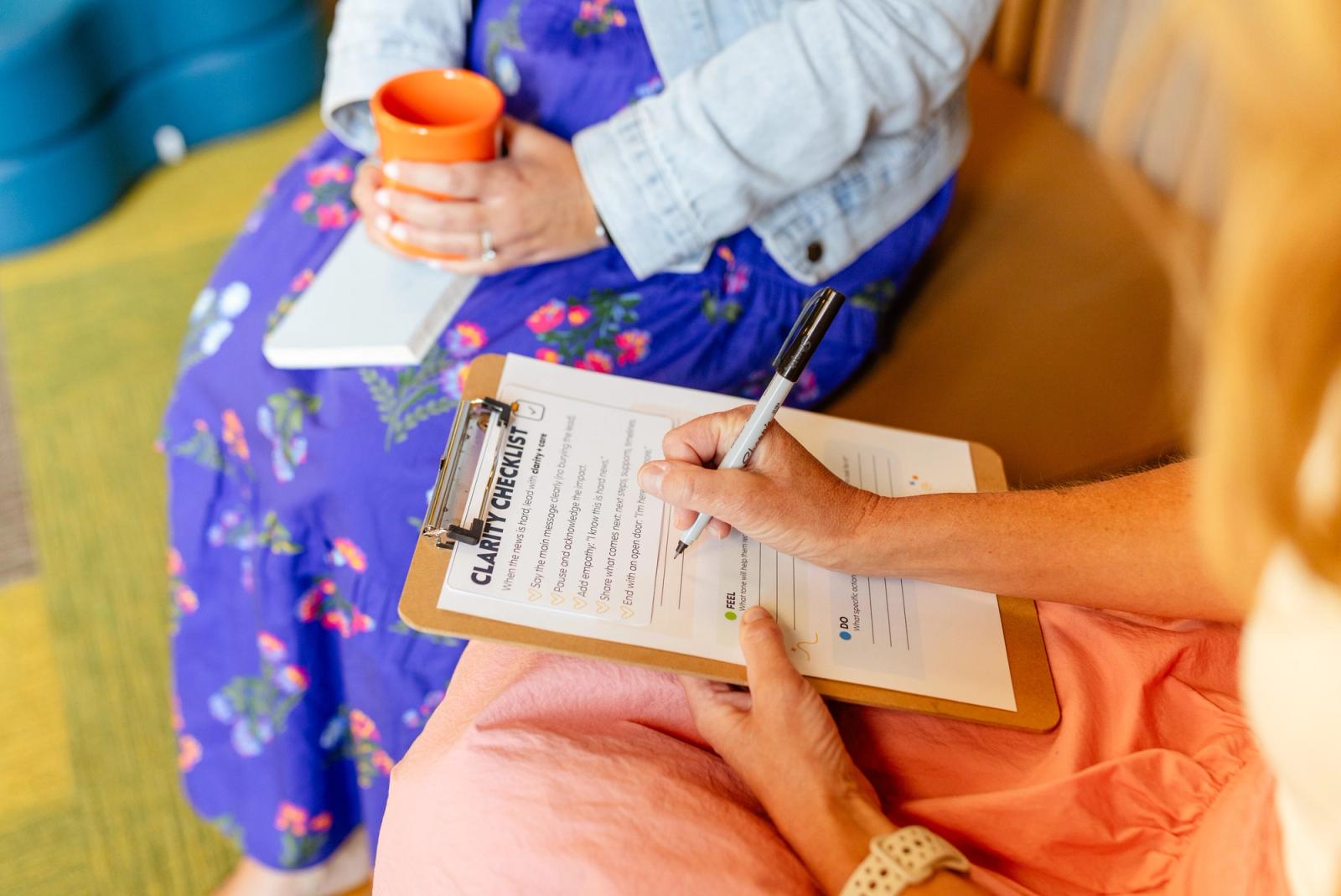 A woman sits on a couch with a clipboard and pen in her lap. Another woman holding a coffee mug sits next to her.