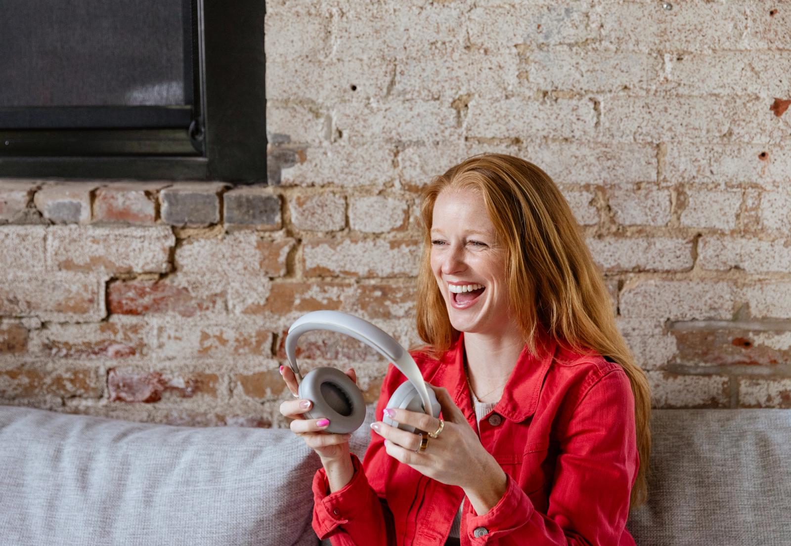 A woman laughs as she places headphones on her hear. She sits in front of a white brick wall.