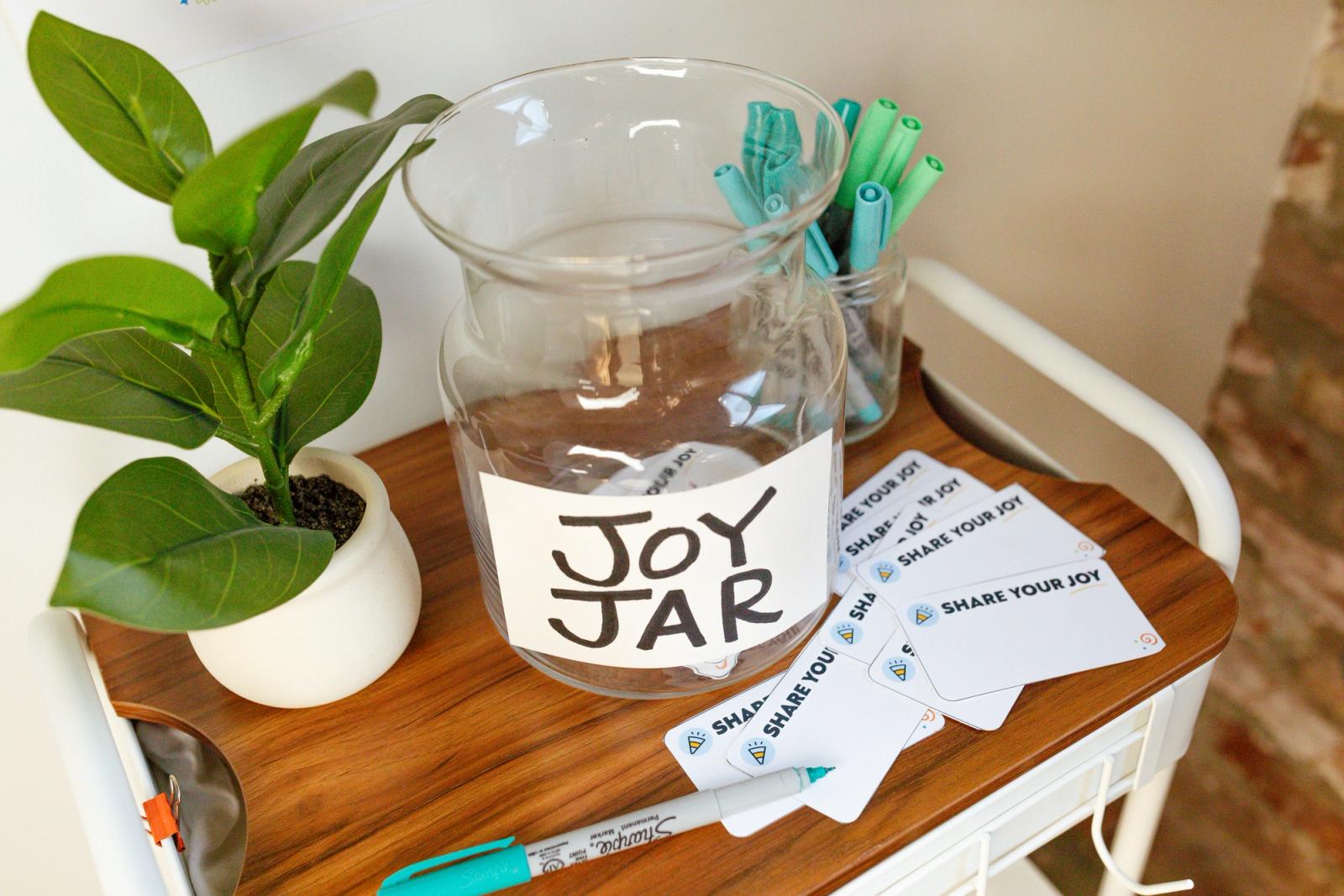 A top view of a cart with a glass jar, potted plant, pens, and notes in front of a white wall.