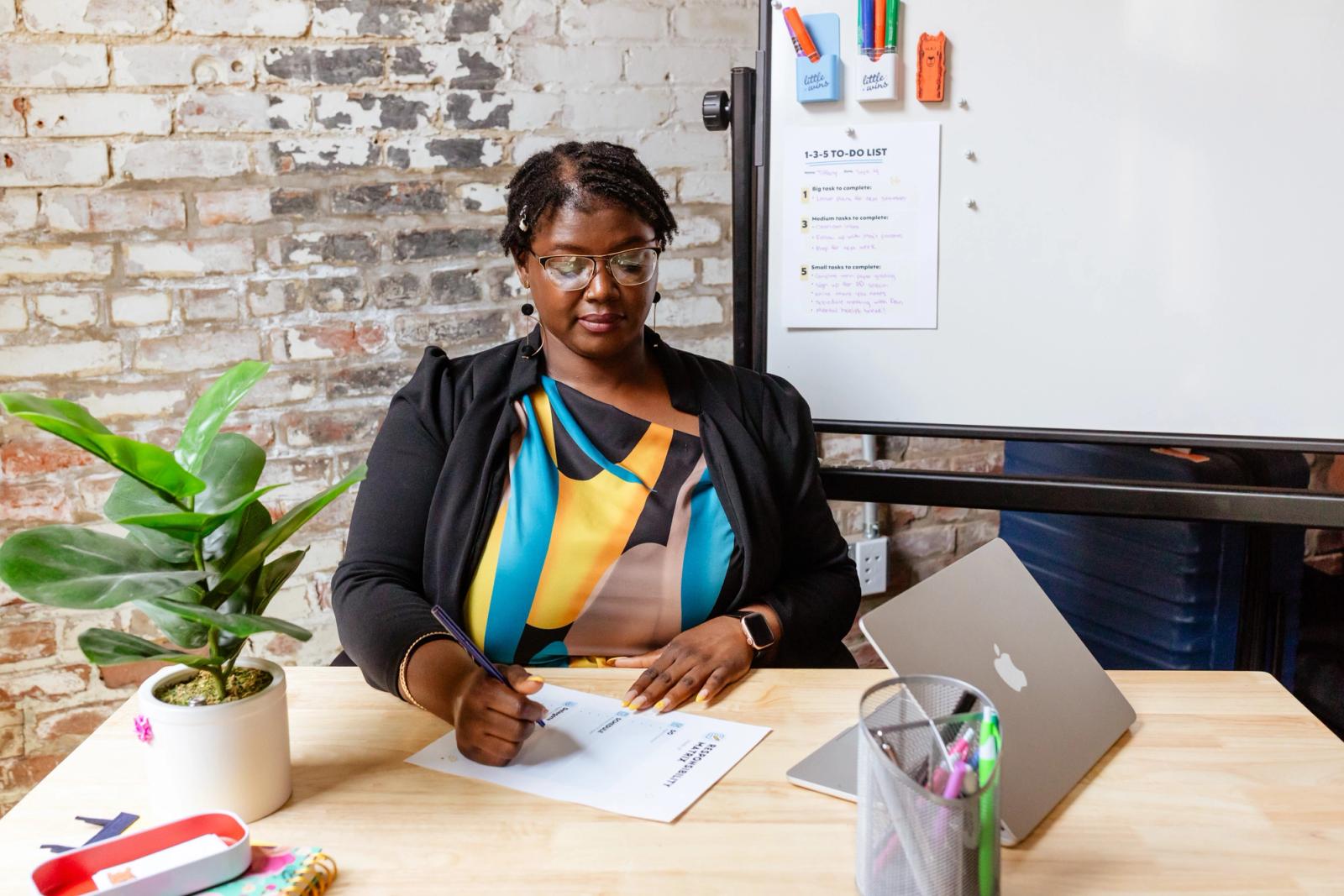 A woman sits at a desk with office supplies on it writing on a piece of paper. In the background is a brick wall and whiteboard with a piece of paper on it.