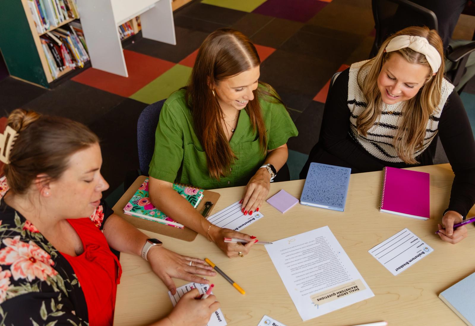 Three women sit at a table in a library. They write on piece of paper and talk with one another. Notebooks, papers, and pens are scattered on the table.