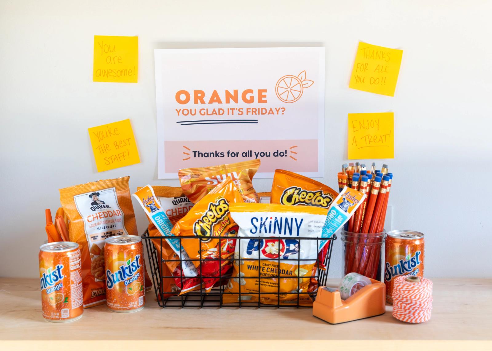 A basket full of orange colored snack bags sits on a desk along with orange office supplies. On the wall is a sign and orange sticky notes with handwritten messages.