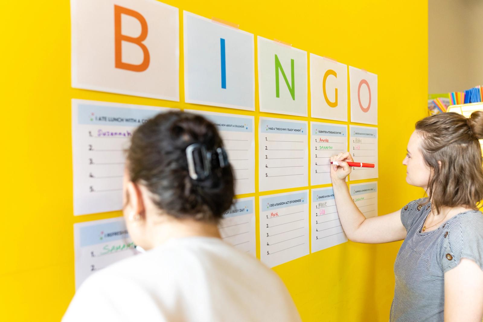 A side view of a yellow wall with sheets of paper that say BINGO on them taped on it. Two women write with markers on the sheets.