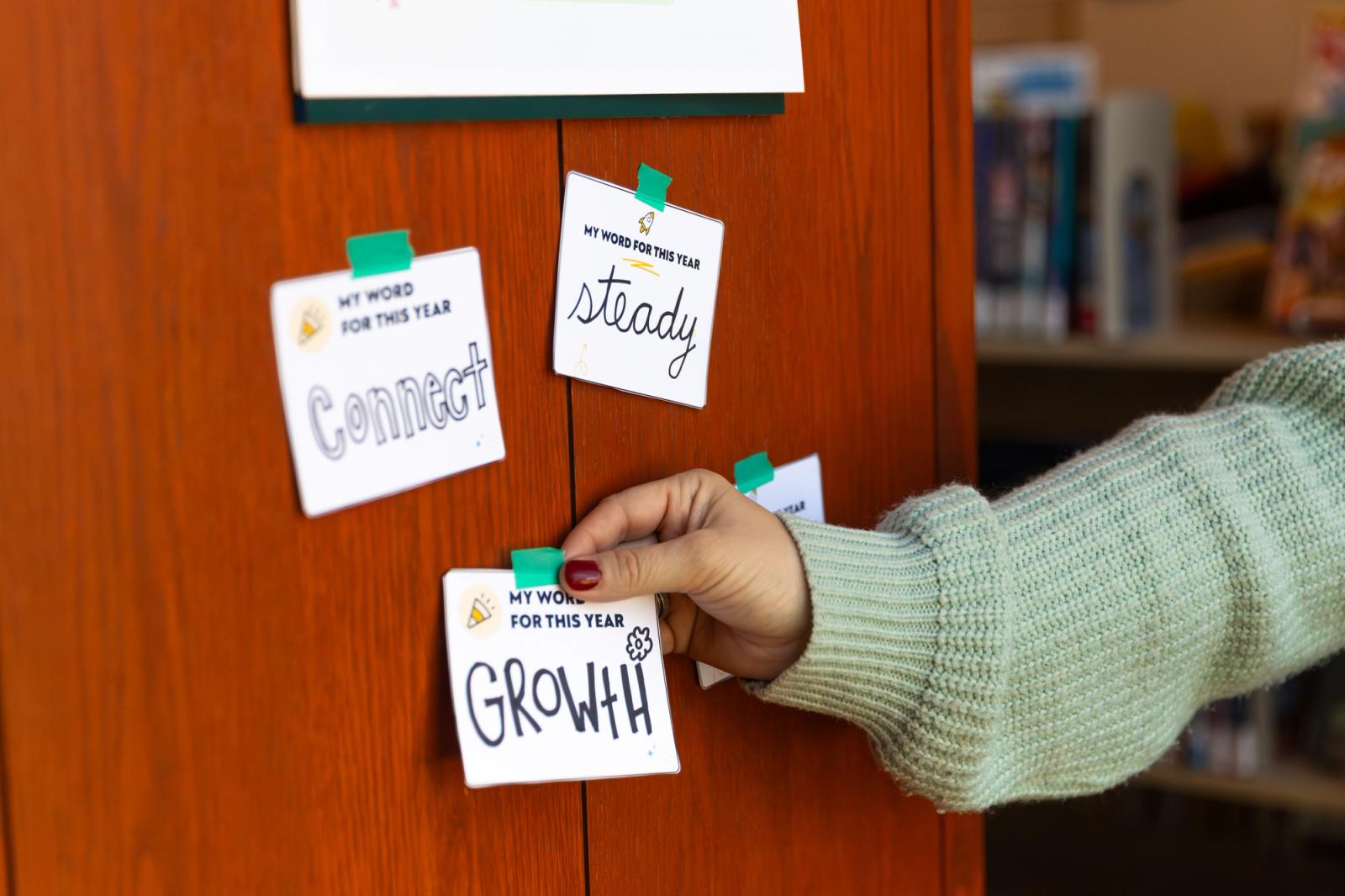 Three notecards each with one word on them are taped to a red wood wall. A hand reaches in from the side to tape one more notecard on the wall.