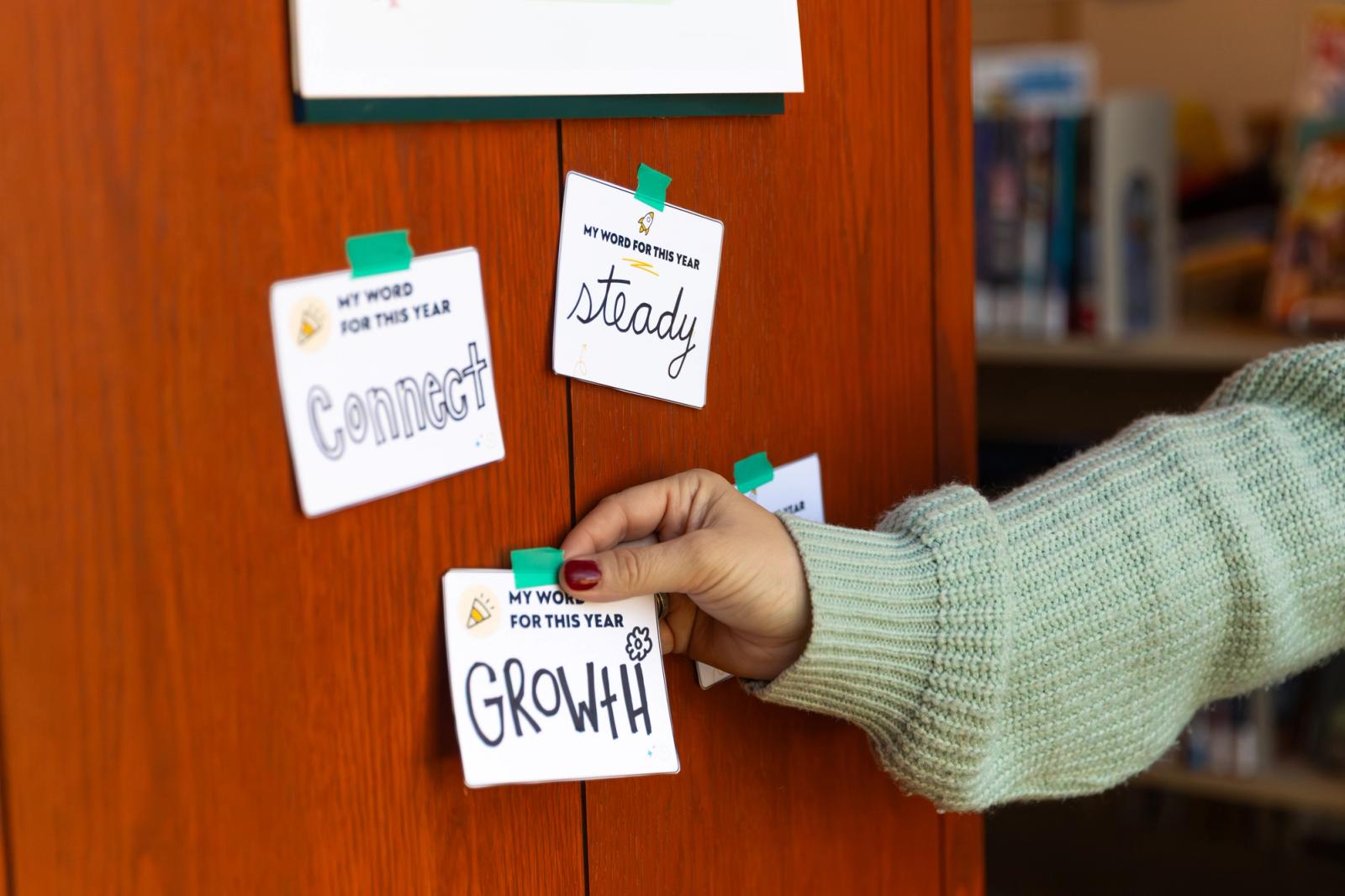 Three notecards each with one word on them are taped to a red wood wall. A hand reaches in from the side to tape one more notecard on the wall.