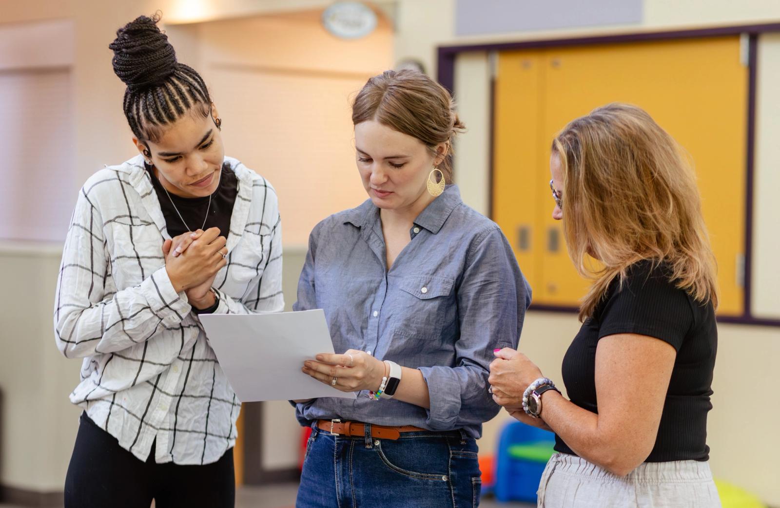 Three women stand in a classroom. The woman in the middle holds a sheet of paper that they all look at.