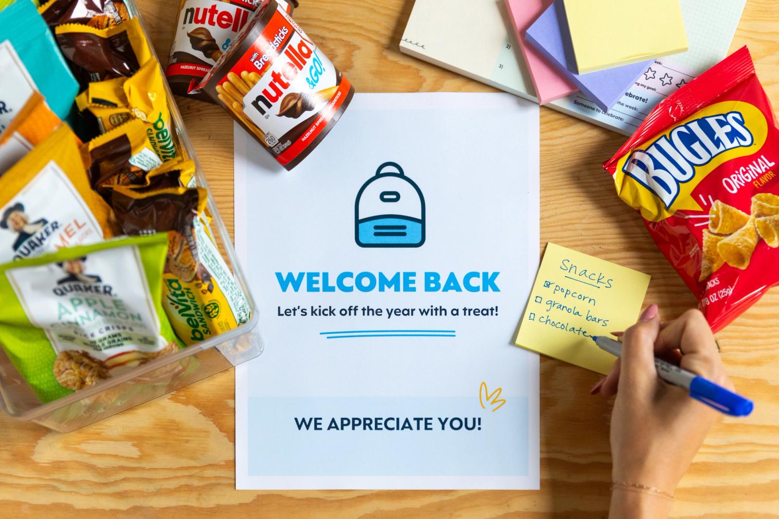 A poster with "welcome back" sits on a wooden table. Next to it is a sticky note pad where someone writes a note with a blue pen. Scattered around are small bags of snacks.