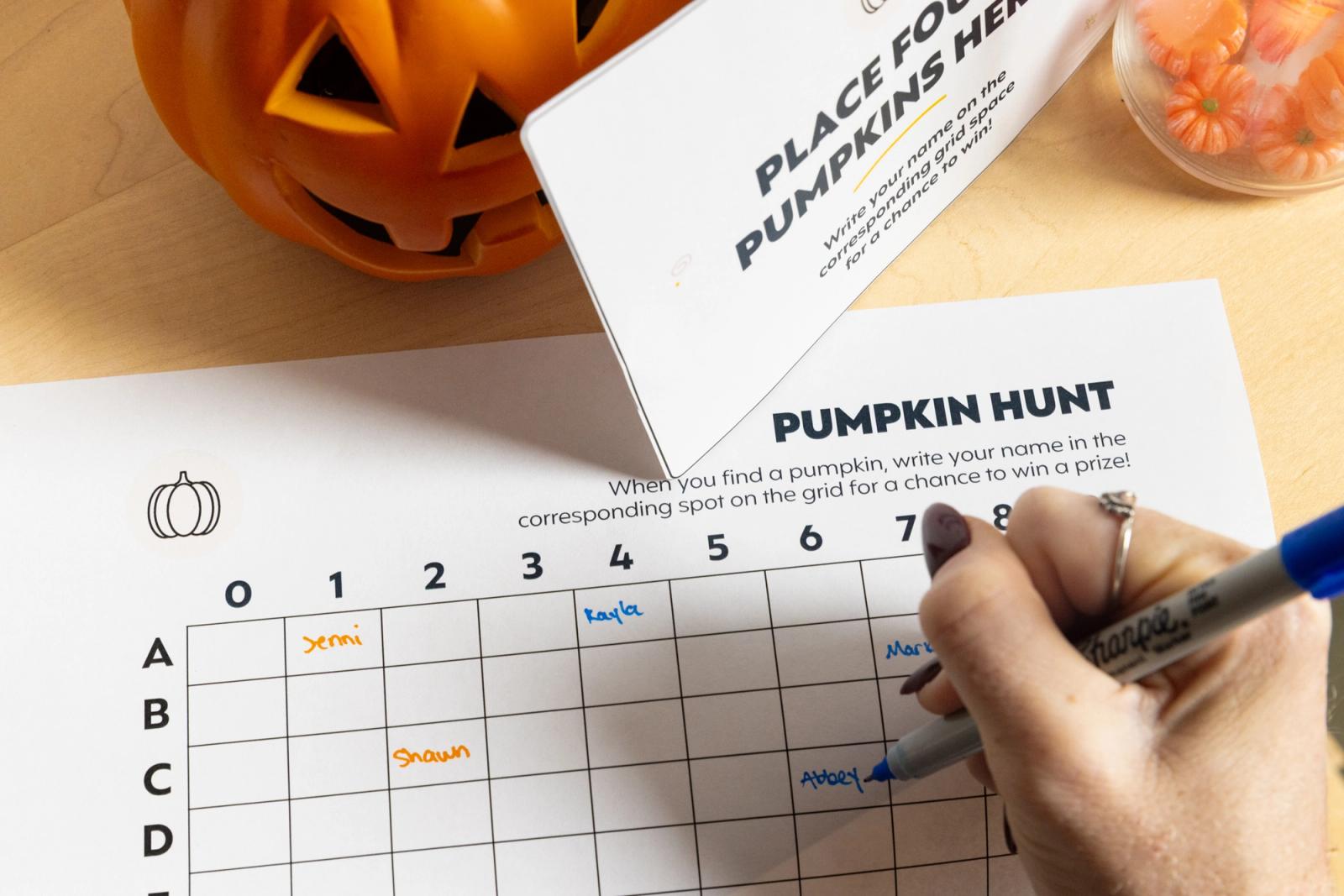 A hand writes on a piece of paper that says "pumpkin hunt" next to a pumpkin.