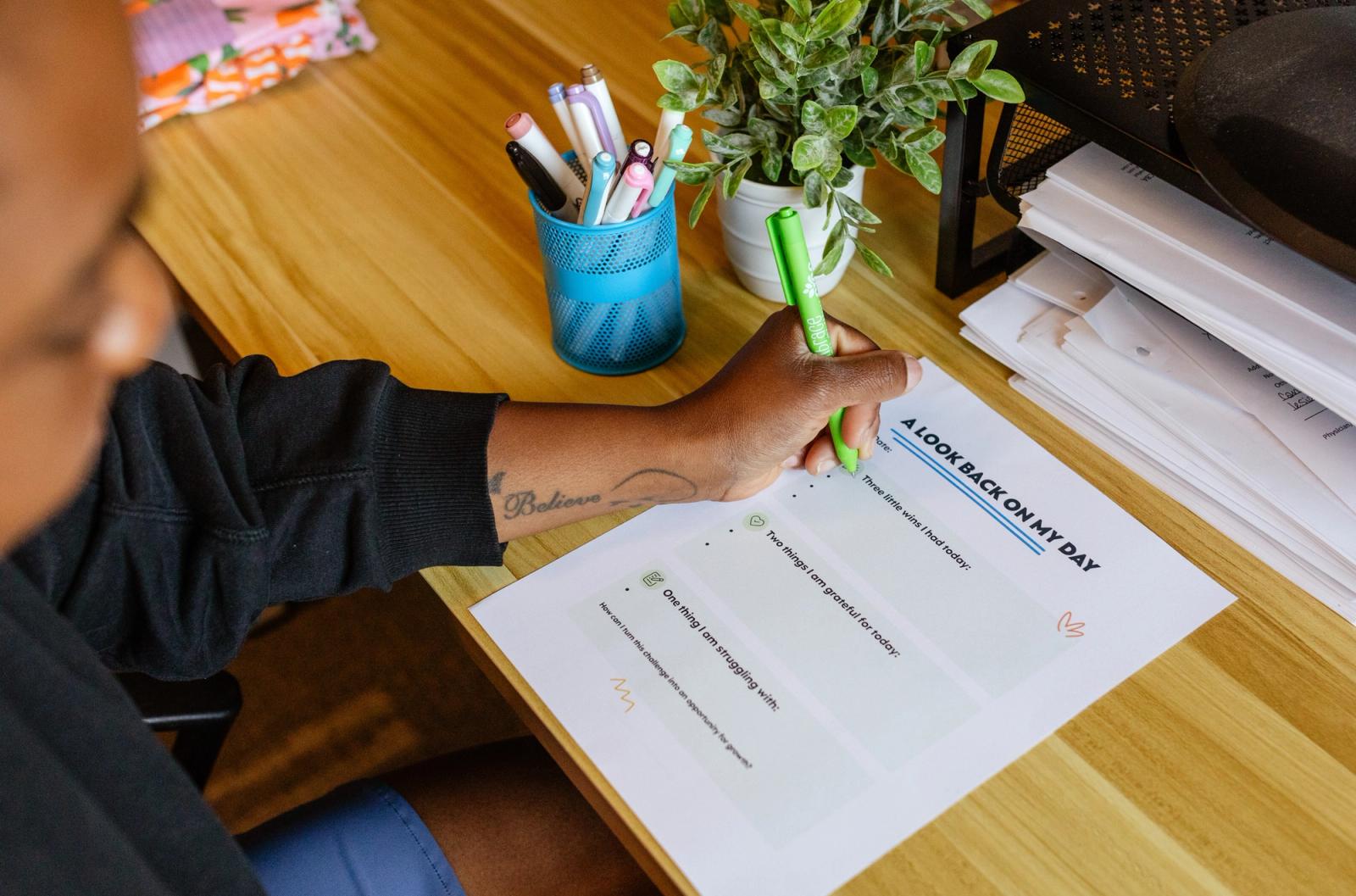 A woman sits at a wooden desk with markers and a potted plant on it. She is writing with a green pen on a sheet of paper.