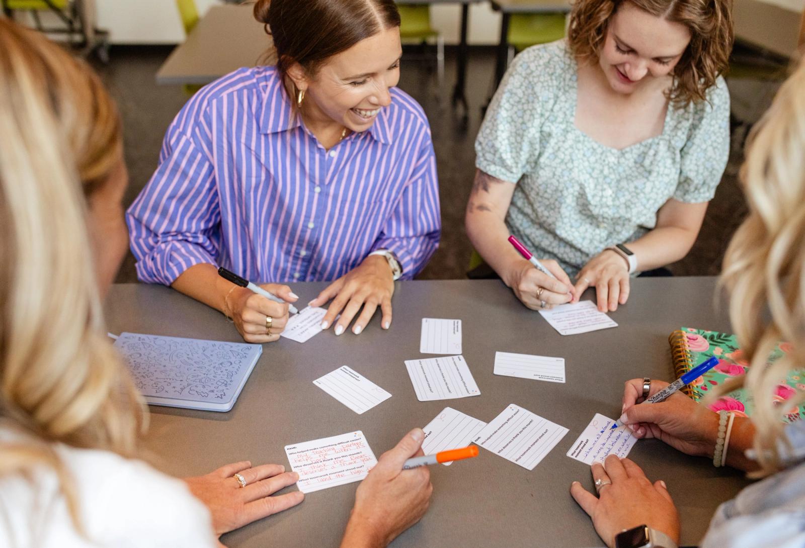Four people sit at a table with notecards scattered on it. One woman looks at another woman writing and smiles. 