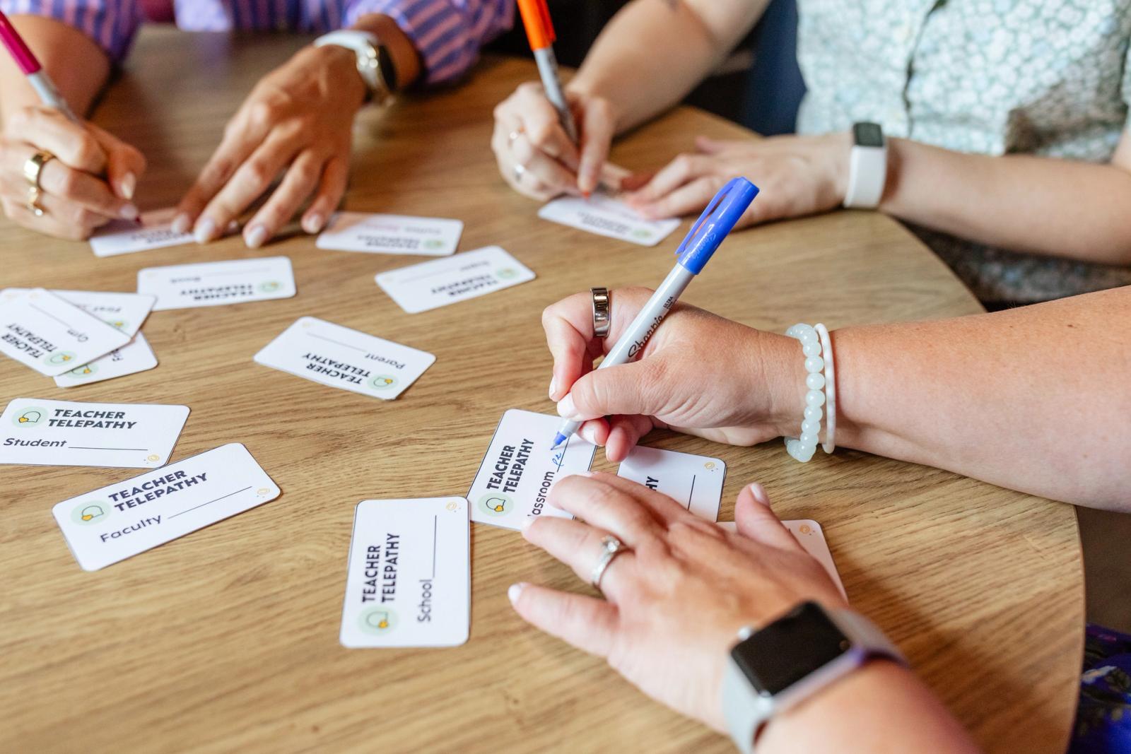 Several small pieces of paper lay on a wood table. Three people sit around the table writing on the pieces of paper.