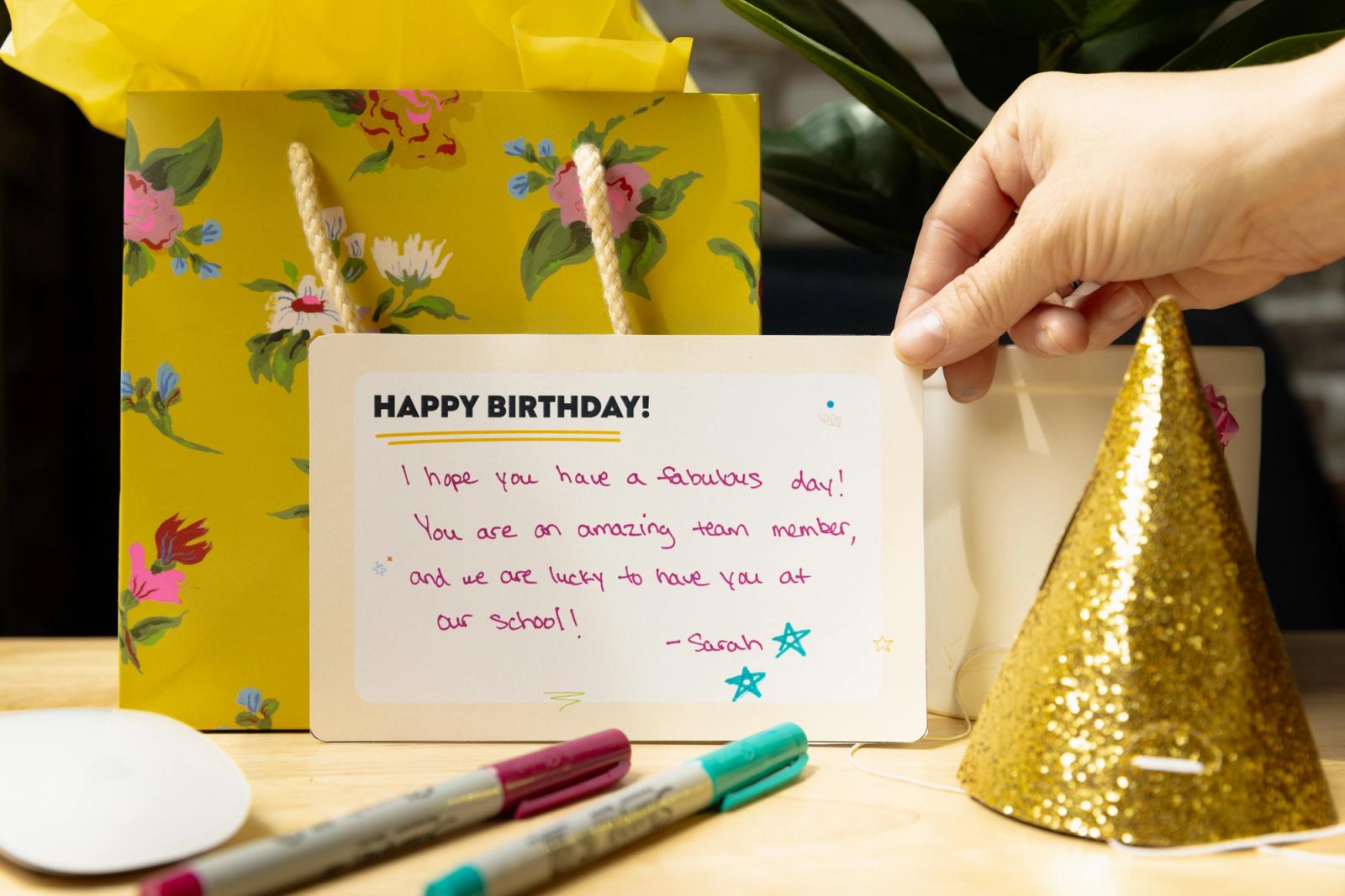 A hand places a note on a desk. Behind it is a gift bag and sparkling birthday hat.