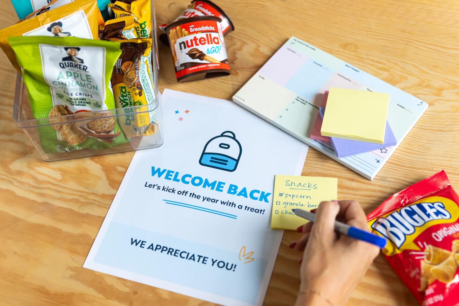 A poster that says "welcome back" lays on a table, snacks lay around it. A hand writes on a sticky note.