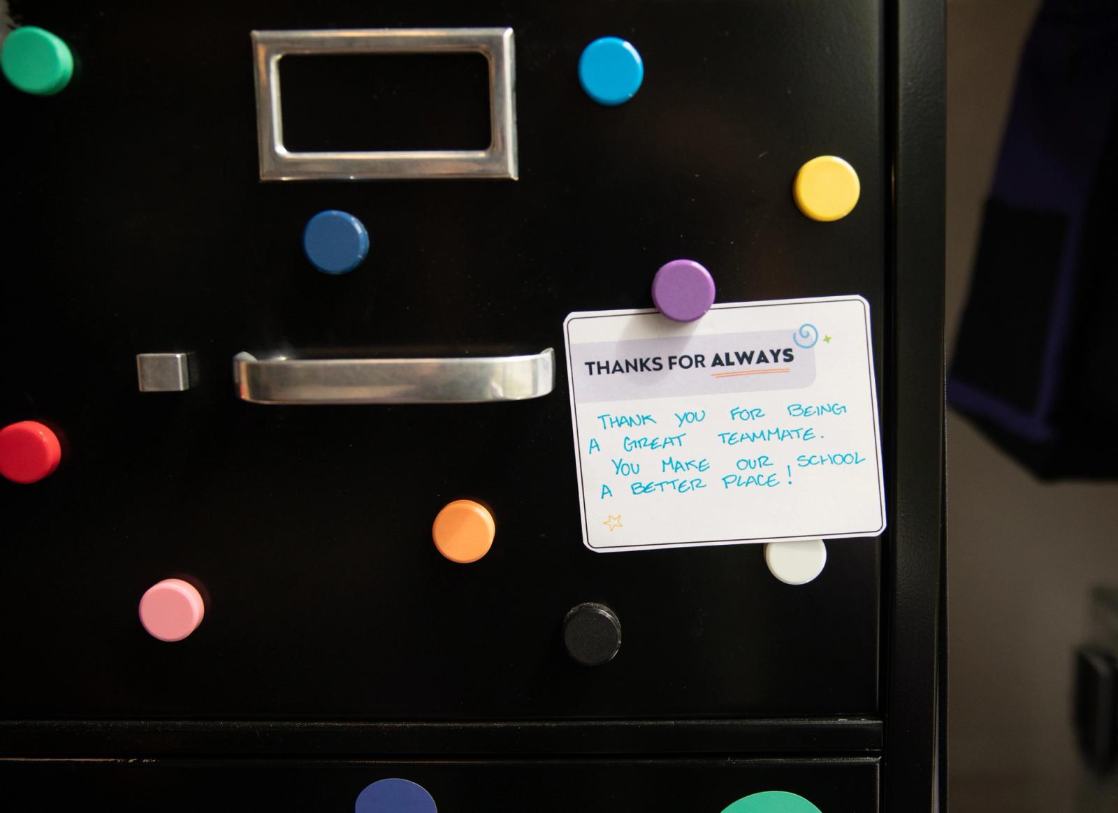 A handwritten note is placed on a black filing cabinet with colorful magnets surrounding it.