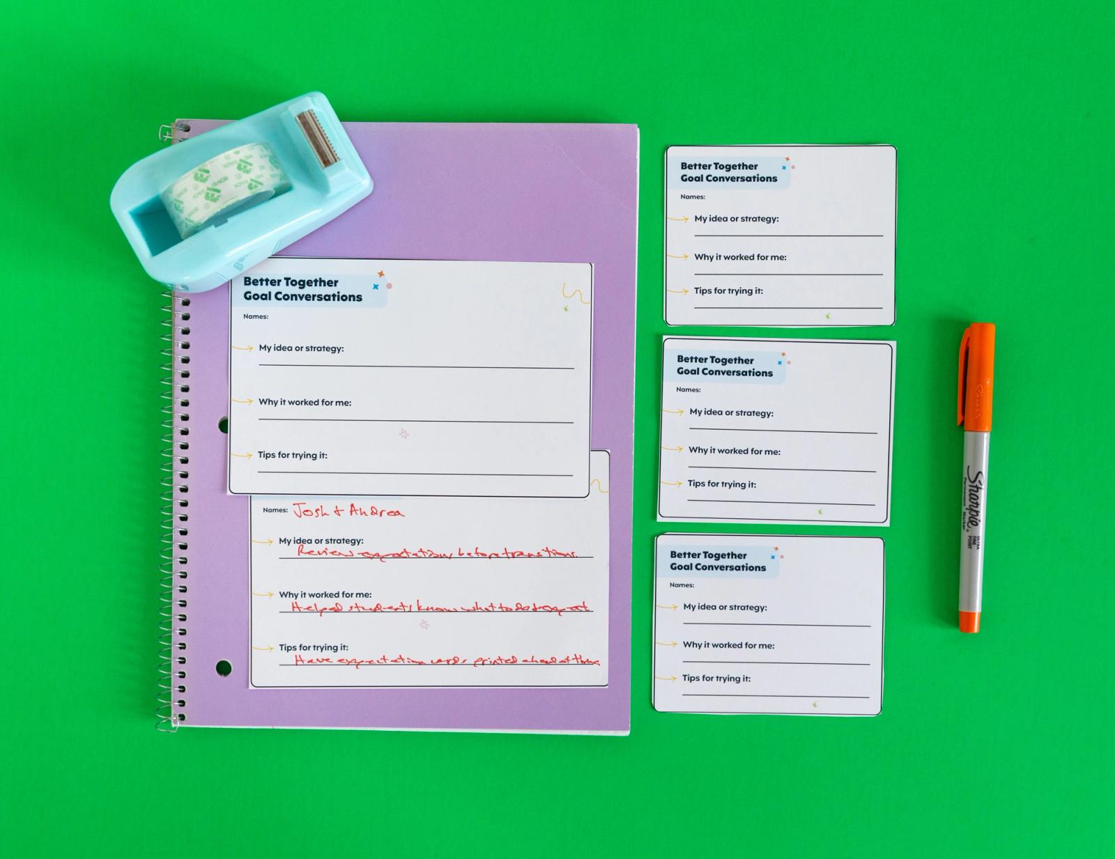 A purple notebook with notecards and a tape dispenser on top of it lay on a green background. Beside it are three notecards and an orange pen.