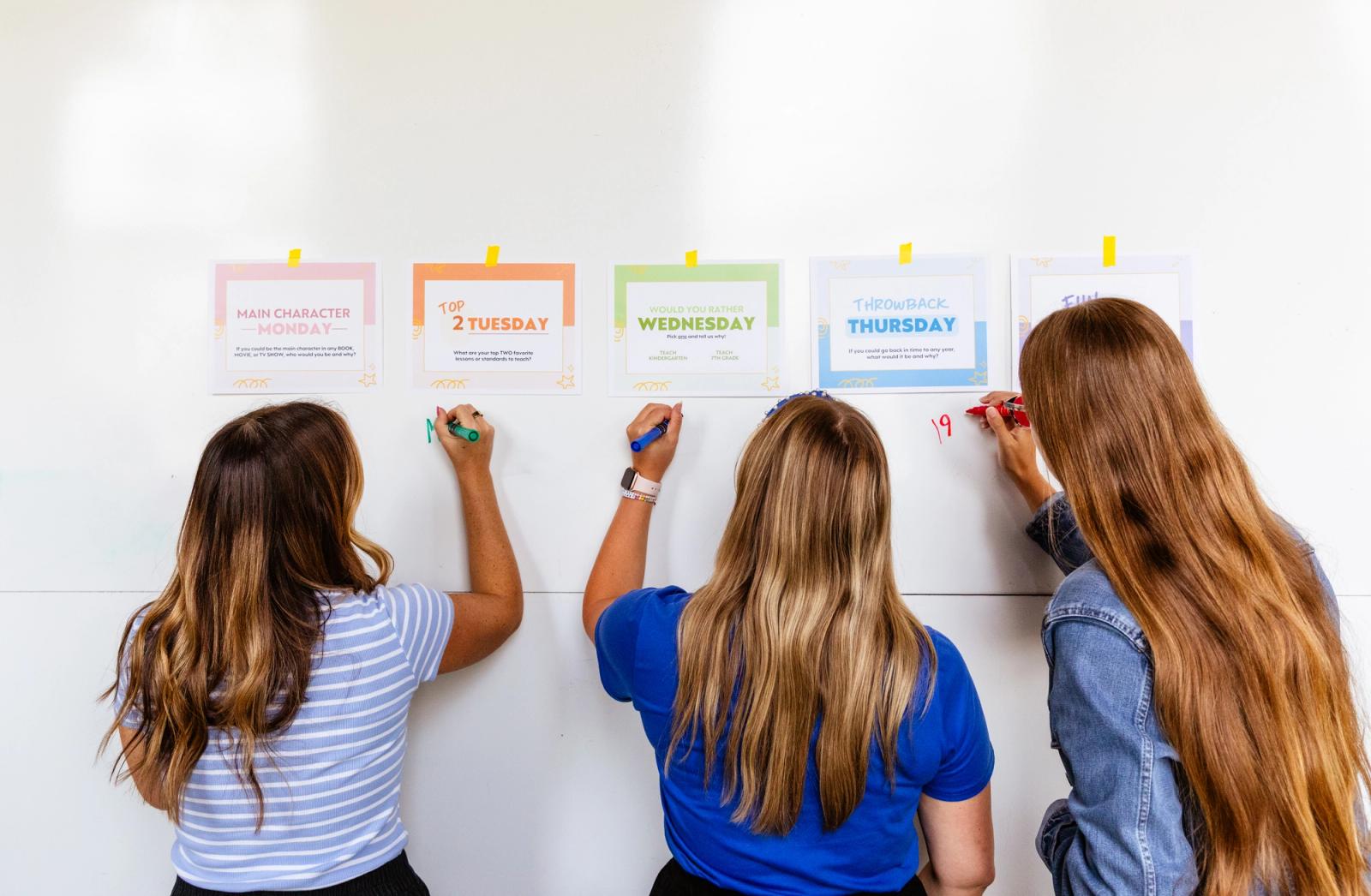 Five pieces of paper are taped to a whiteboard. Three women stand in front of it writing underneath the papers.