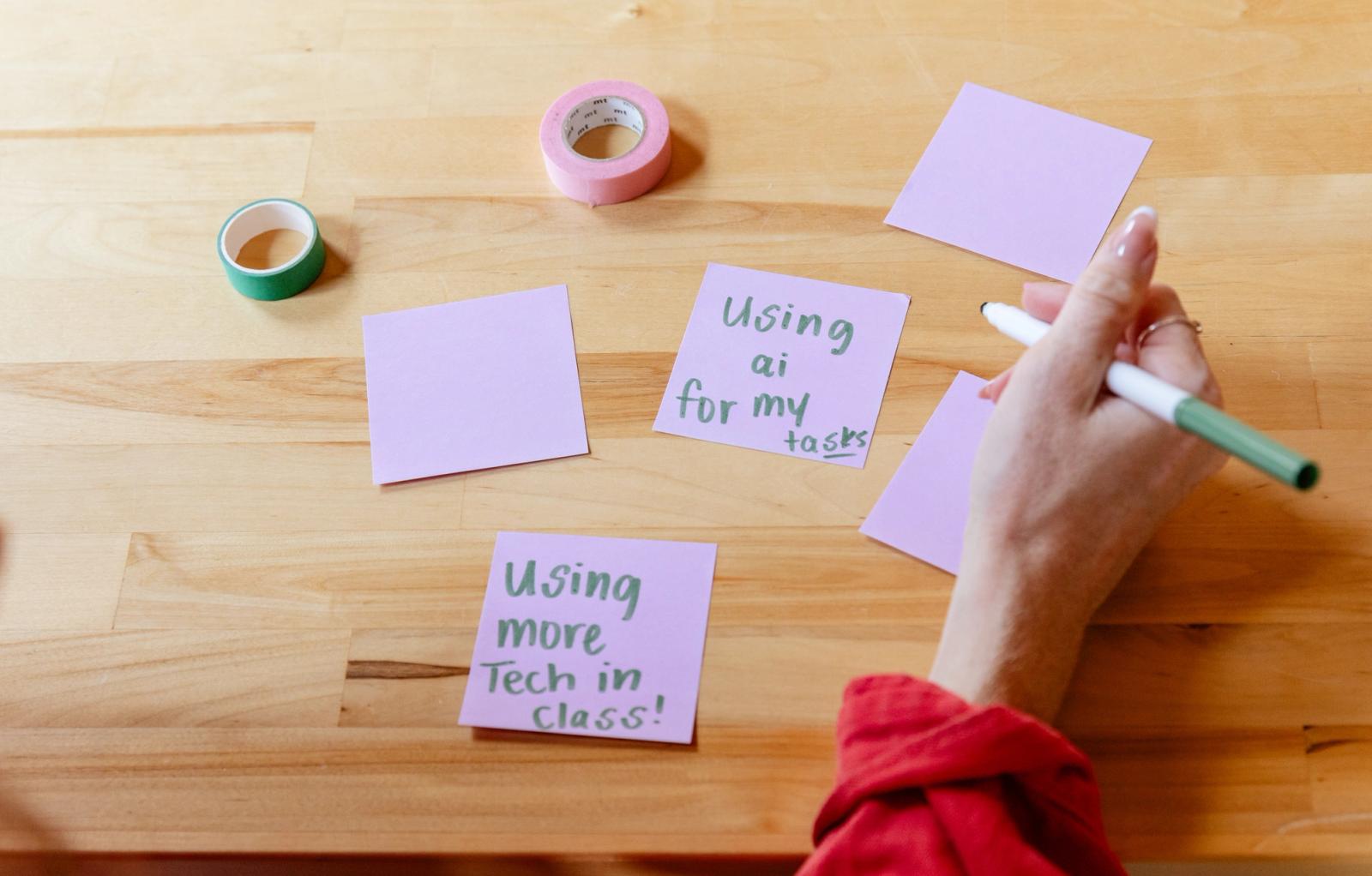 Purple sticky notes with writing on them lay on a wood table. A hand holds a marker to write on another note.