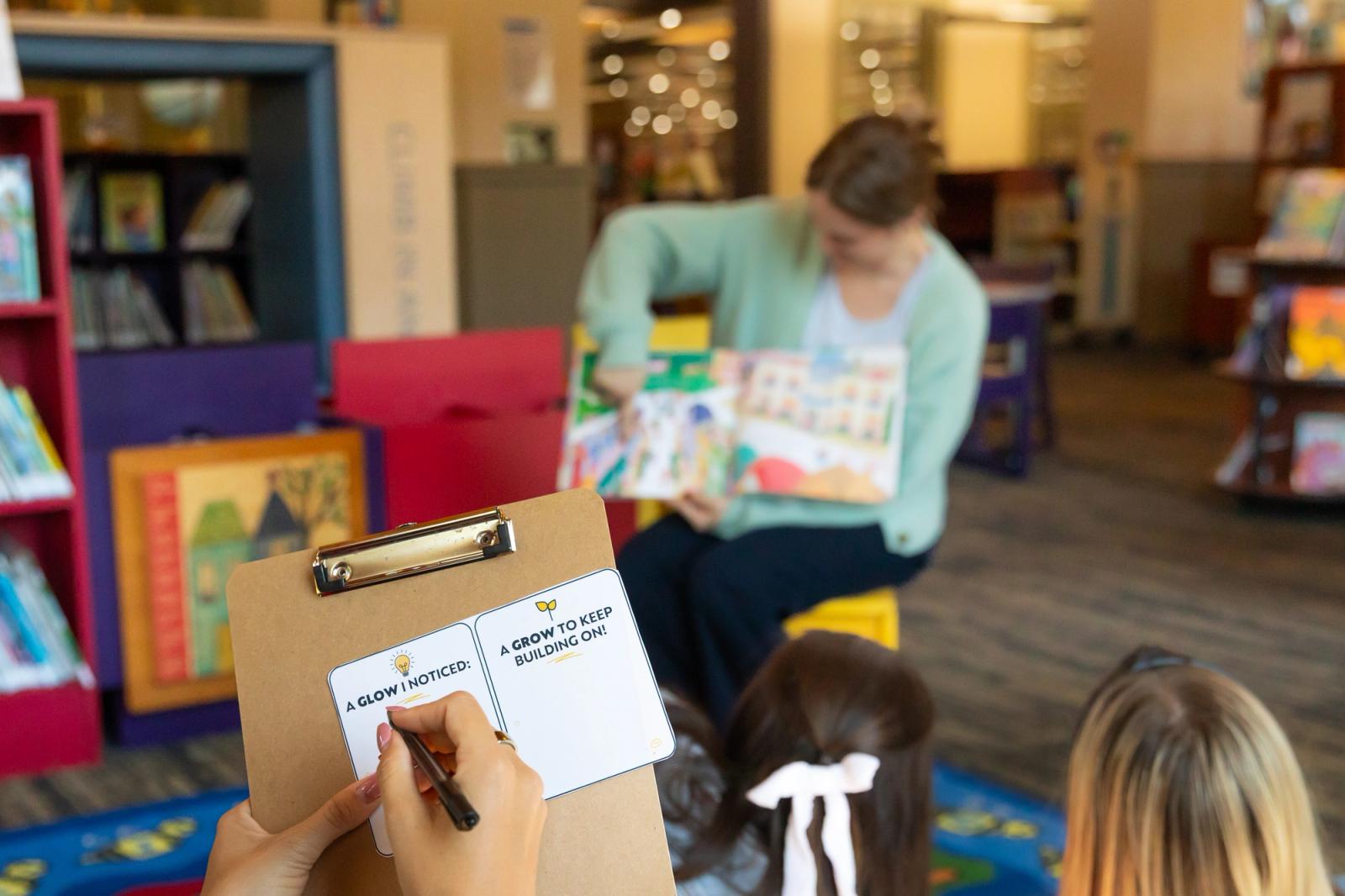 A woman sits in a chair reading a book to a class. Another women faces her and fills out a notecard.