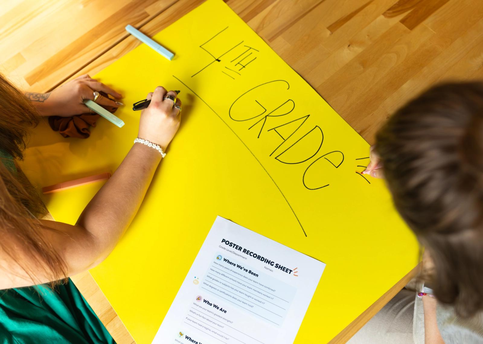 Two women sit at a table with a large yellow posterboard on it. There is a sheet of paper and markers, and the women are writing on the poster.