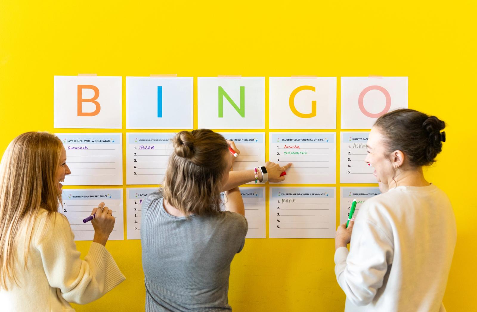 Three people stand in front of a yellow wall with pens in their hands, pointing and laughing. Sheets of paper that say BINGO are taped on the wall.
